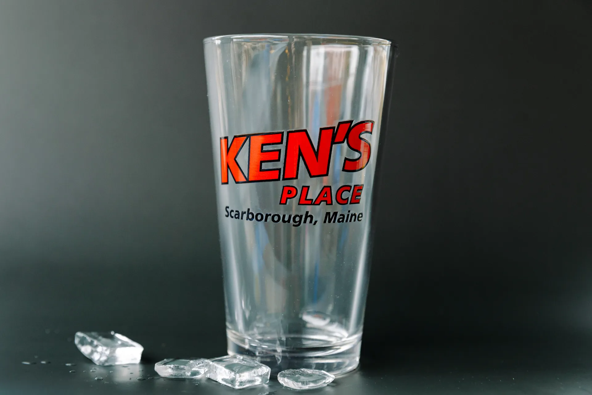Clear drinking glass with red “KEN’S PLACE” logo on a dark background, beside a small ice cube