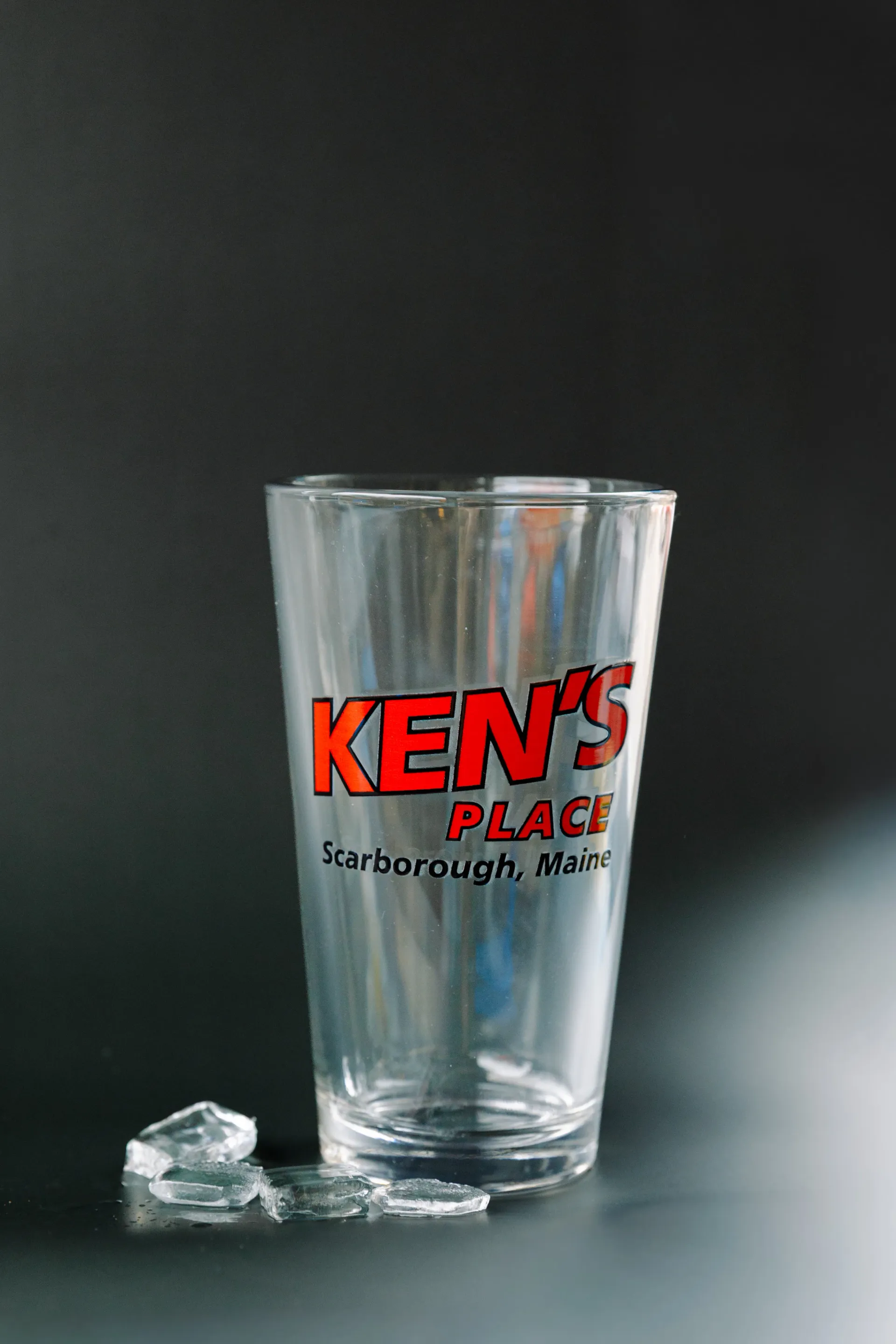 Clear drinking glass with red “KEN’S PLACE” logo on a dark background, beside a small ice cube