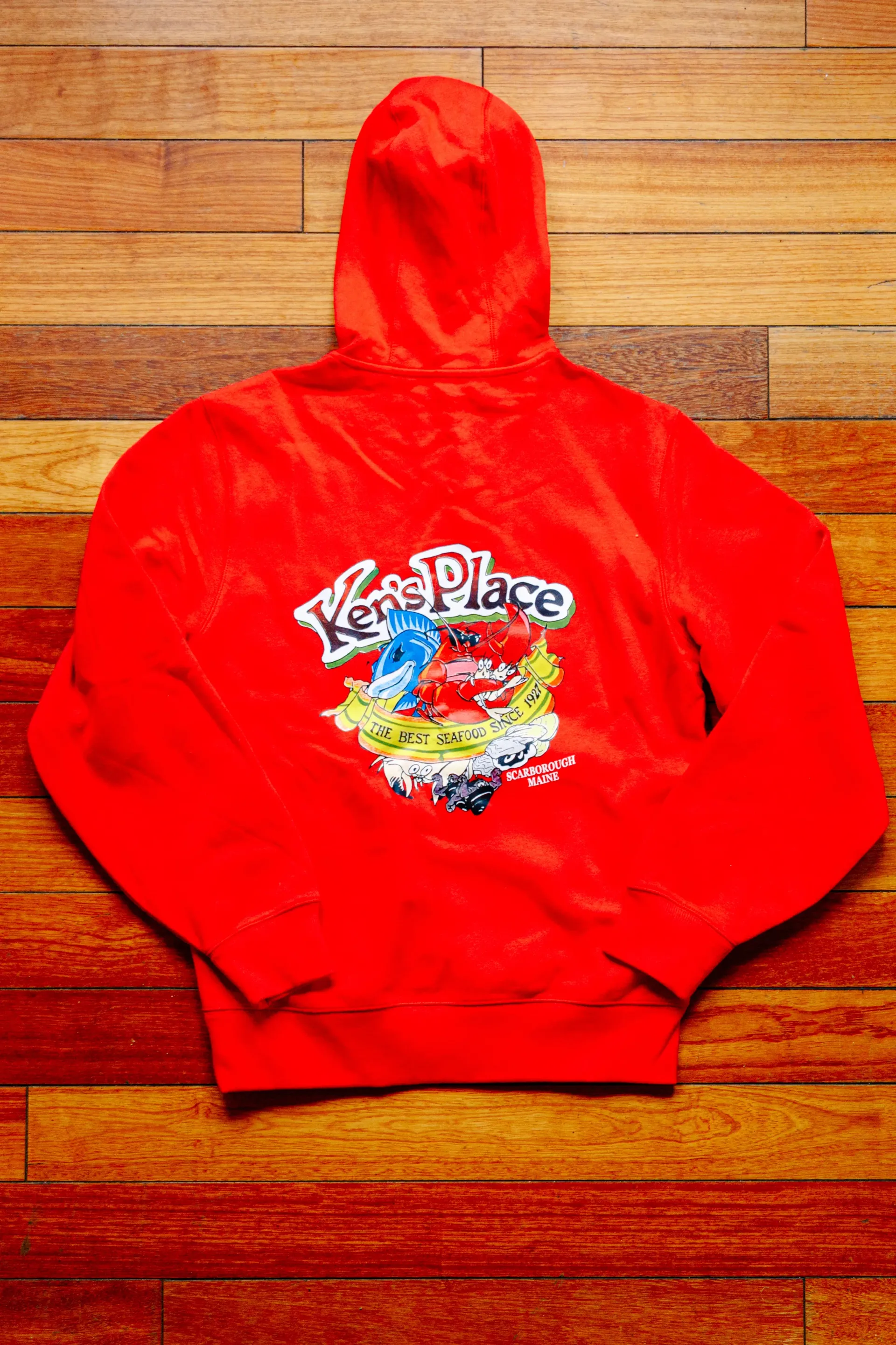 Red hoodie with colorful graphic logo laid flat on a wooden floor.