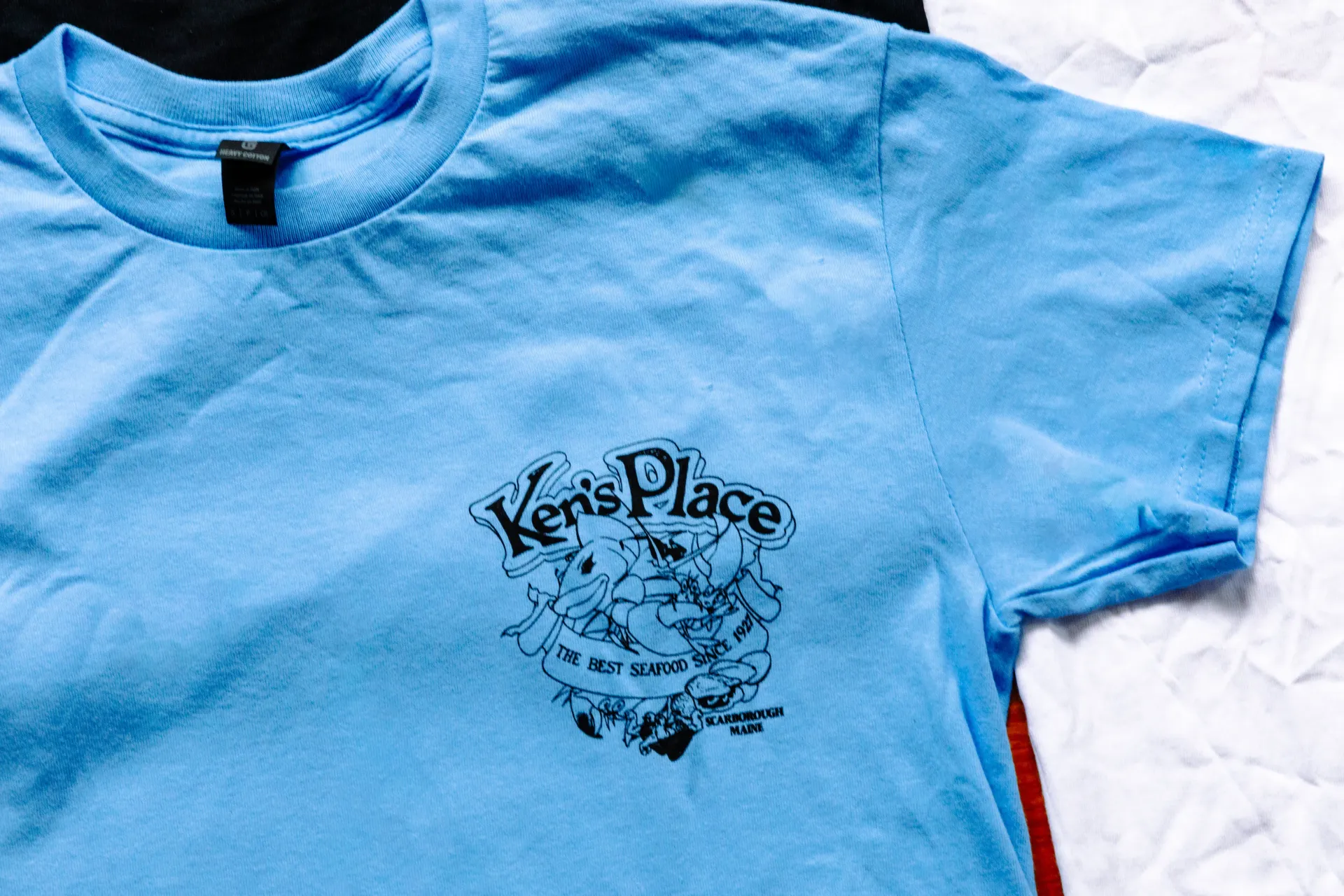 Black T-shirt with colorful graphic and “Kern Place” text, laid on a bed with red, gray, and white bedding