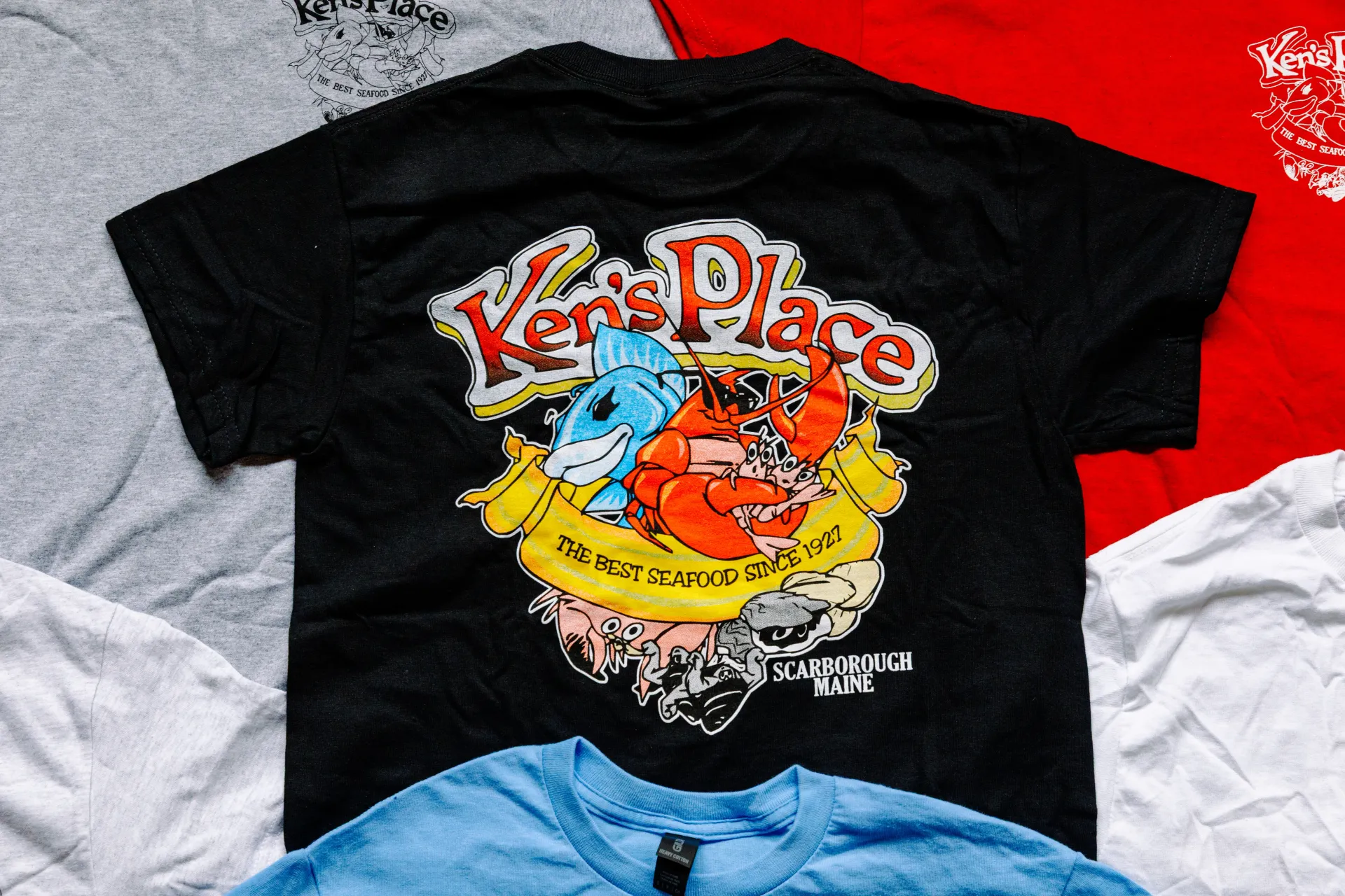 Black T-shirt with colorful graphic and “Kern Place” text, laid on a bed with red, gray, and white bedding
