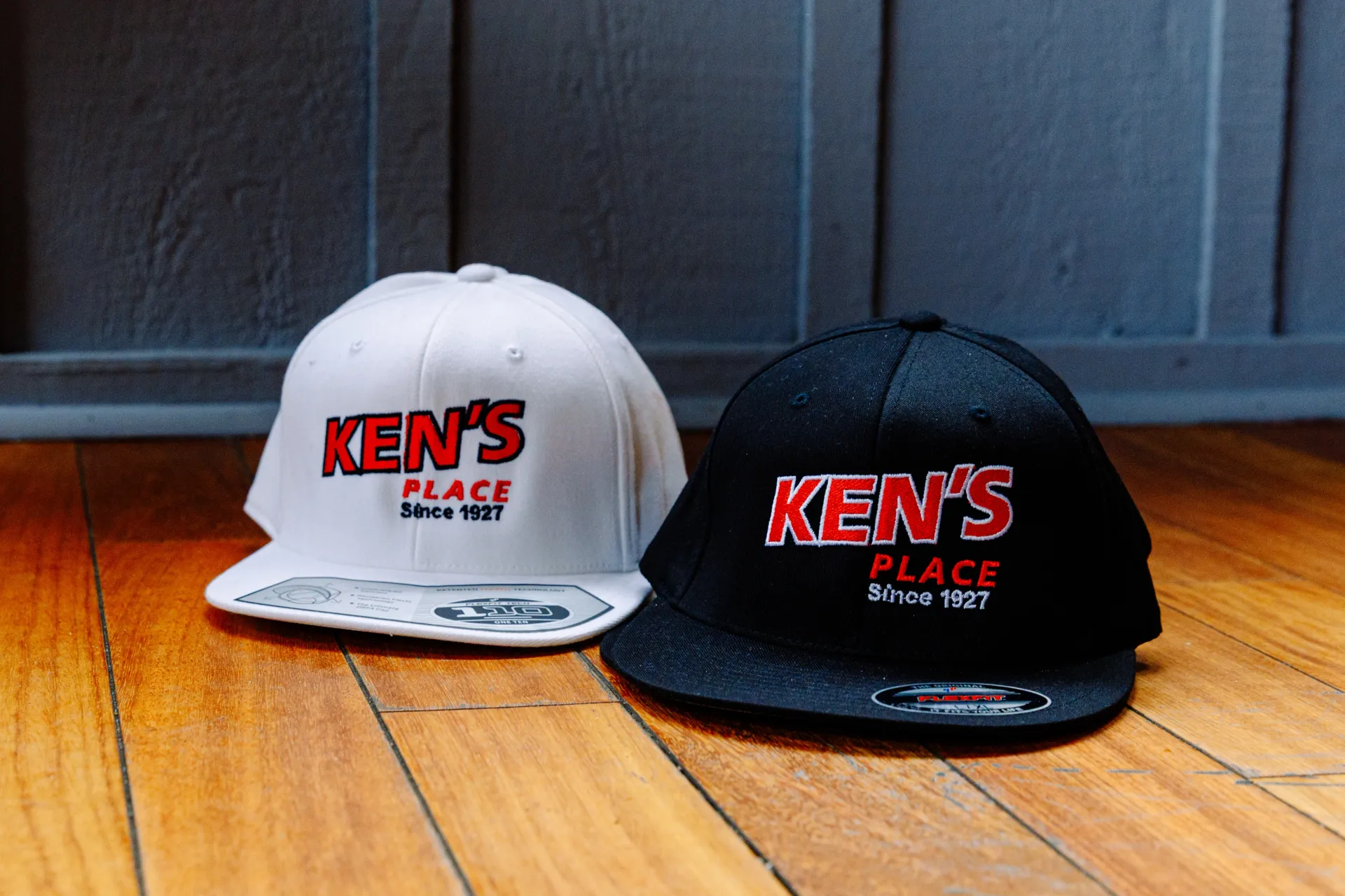 Two Kens Place hats, one white and one black, sitting on a wooden floor against a dark wall.