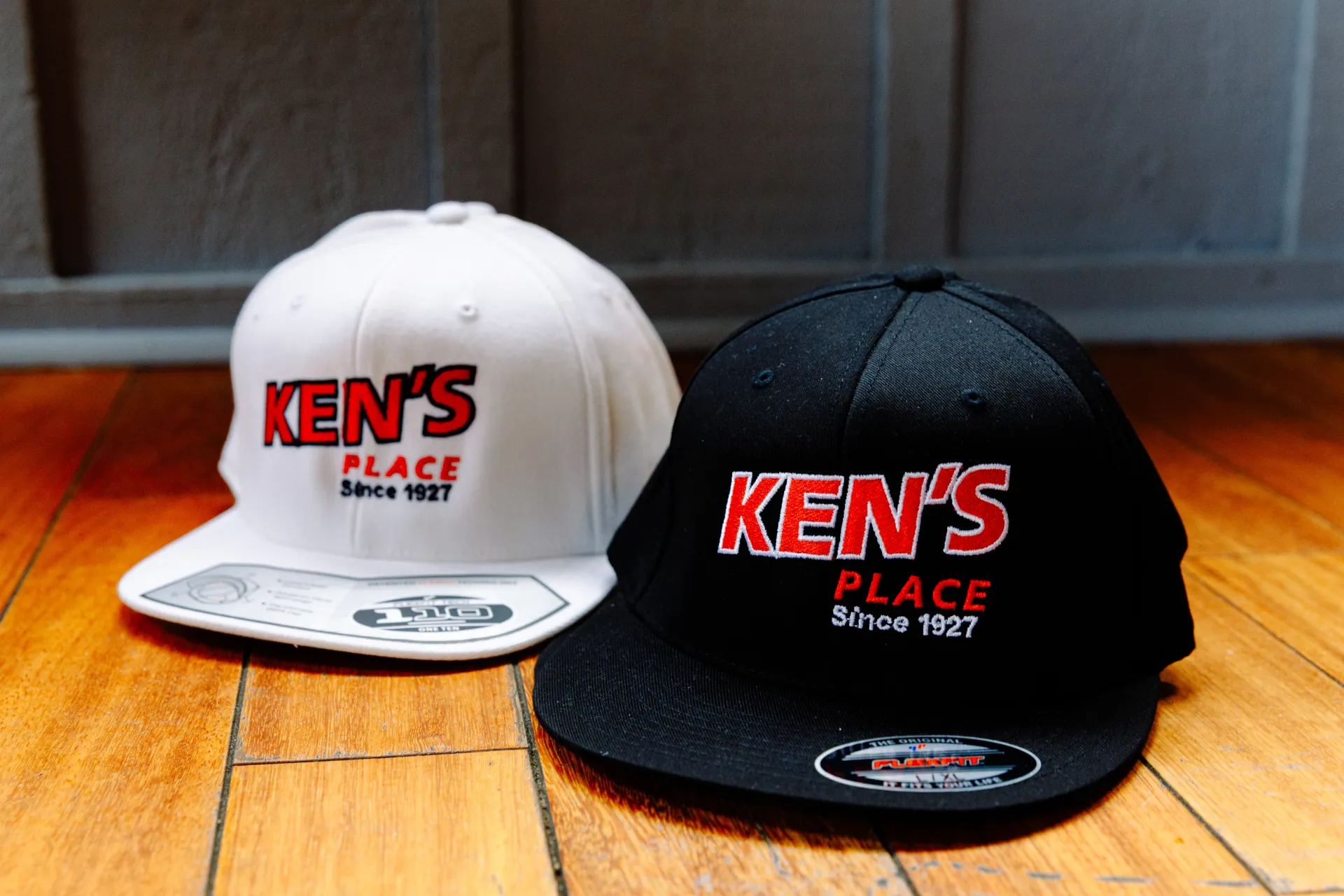 Two Kens Place hats, one white and one black, sitting on a wooden floor against a dark wall.