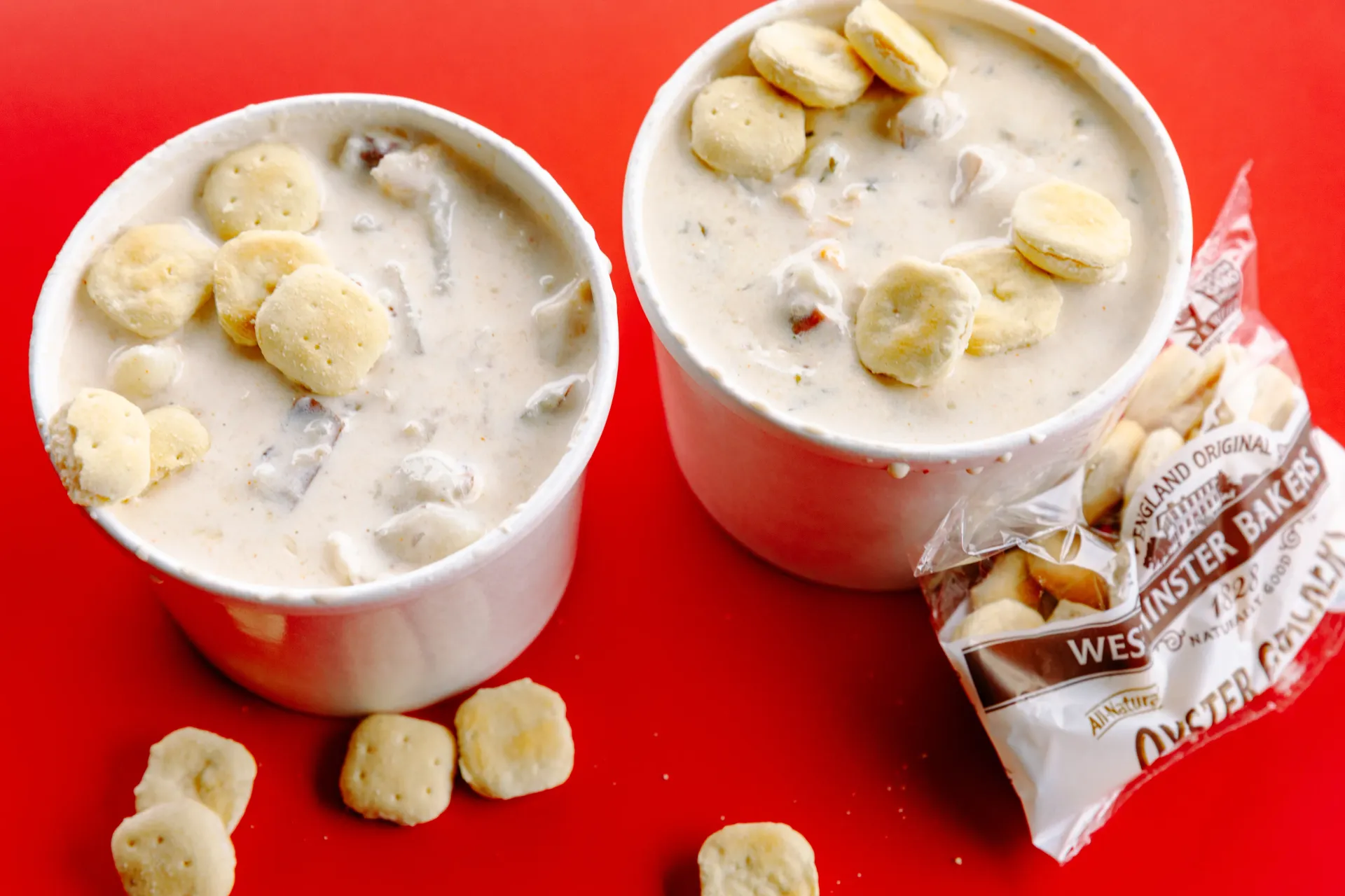 Two cups of creamy dessert topped with banana pieces on a red surface, with a wrapped wafer beside them