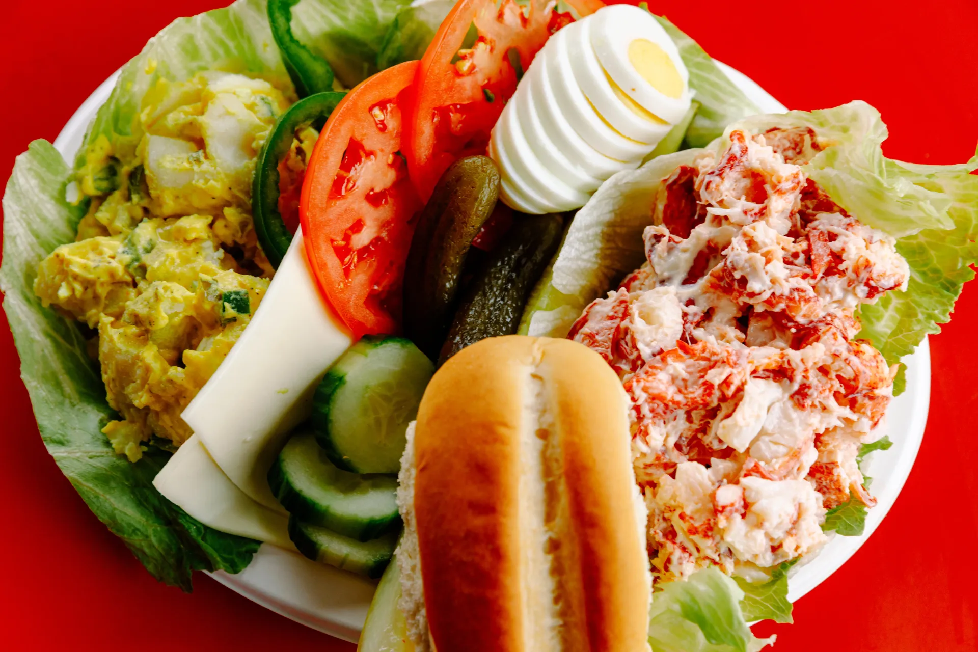 Vegetable platter with lettuce, tomato, cucumber, pickles, boiled egg, tuna salad, and a bread roll on a white plate