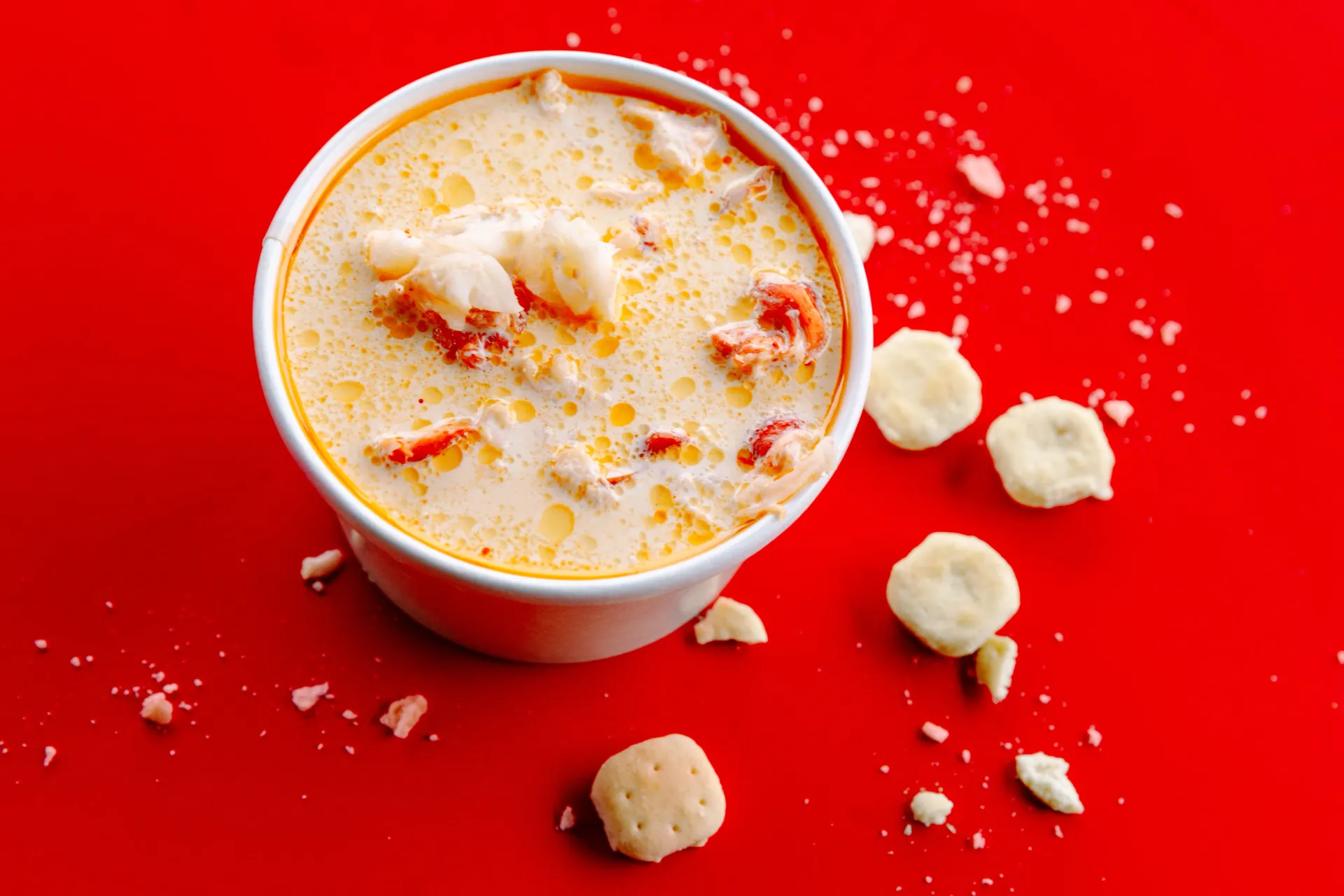Cup of creamy dessert with fruit pieces on a bright red background, with scattered crumbs and small scoops nearby
