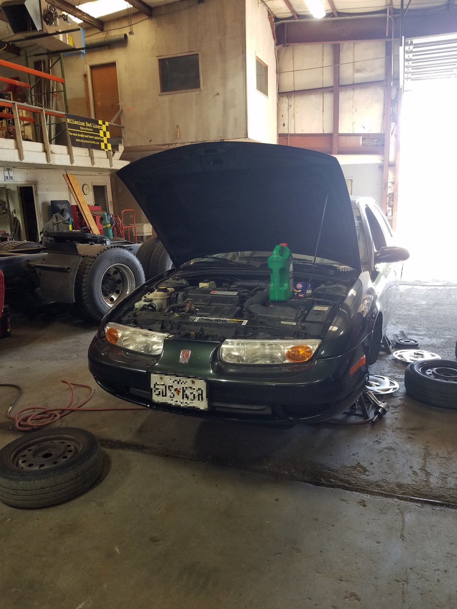 A dark green sedan with its hood open, parked in a cluttered garage with a spare tire on the floor in the foreground.