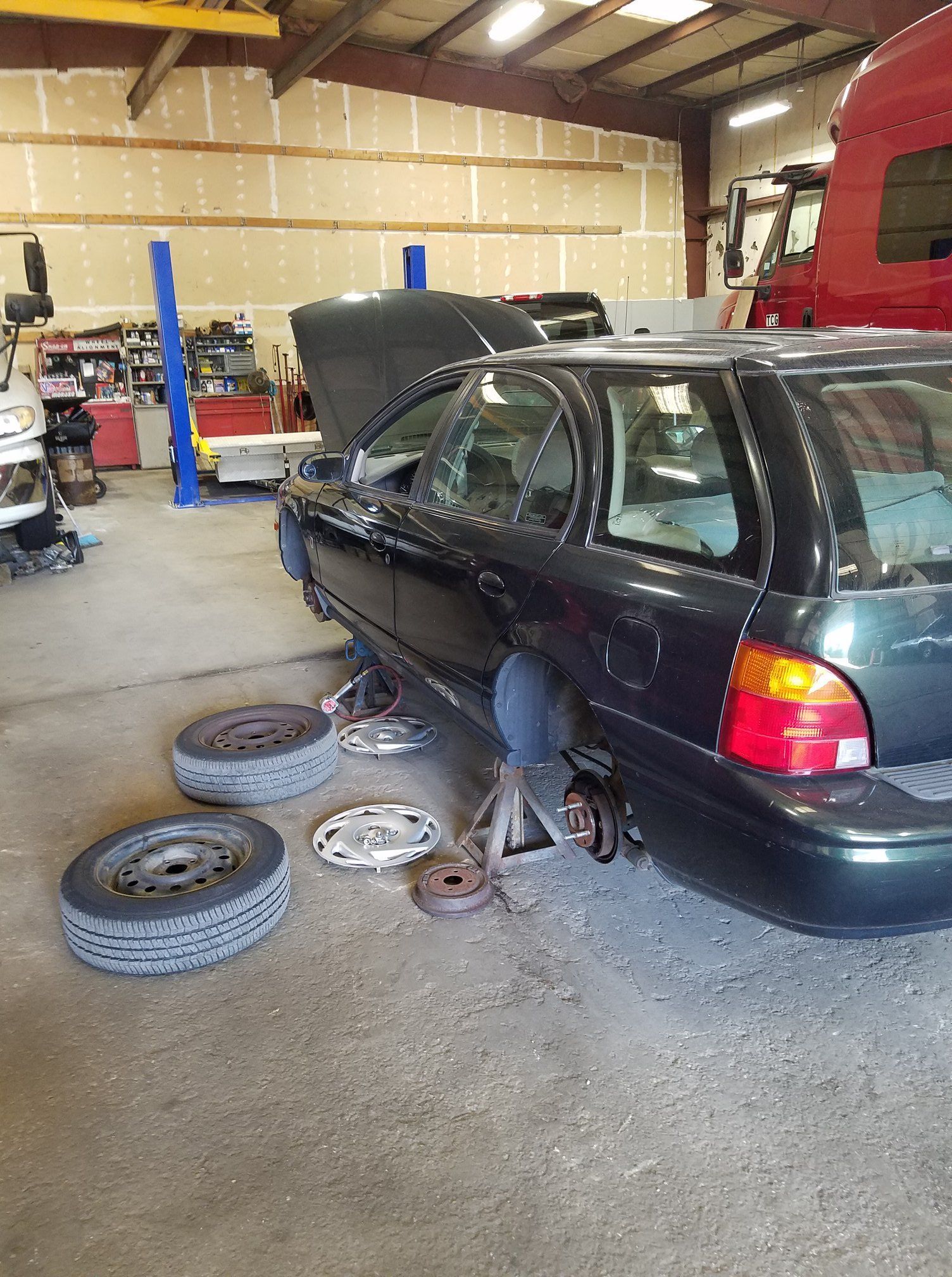 A black station wagon is raised on a jack stand in an auto repair shop with two wheels removed on the garage floor.
