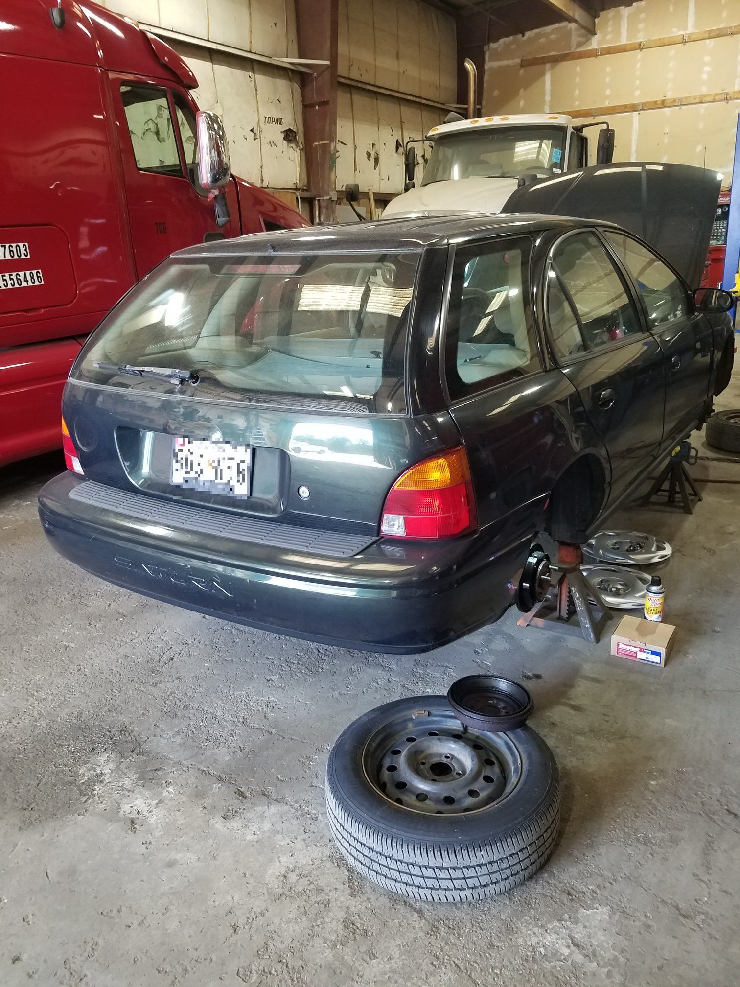 A dark green station wagon sits on jack stands in an auto repair shop with a detached tire resting on the floor.