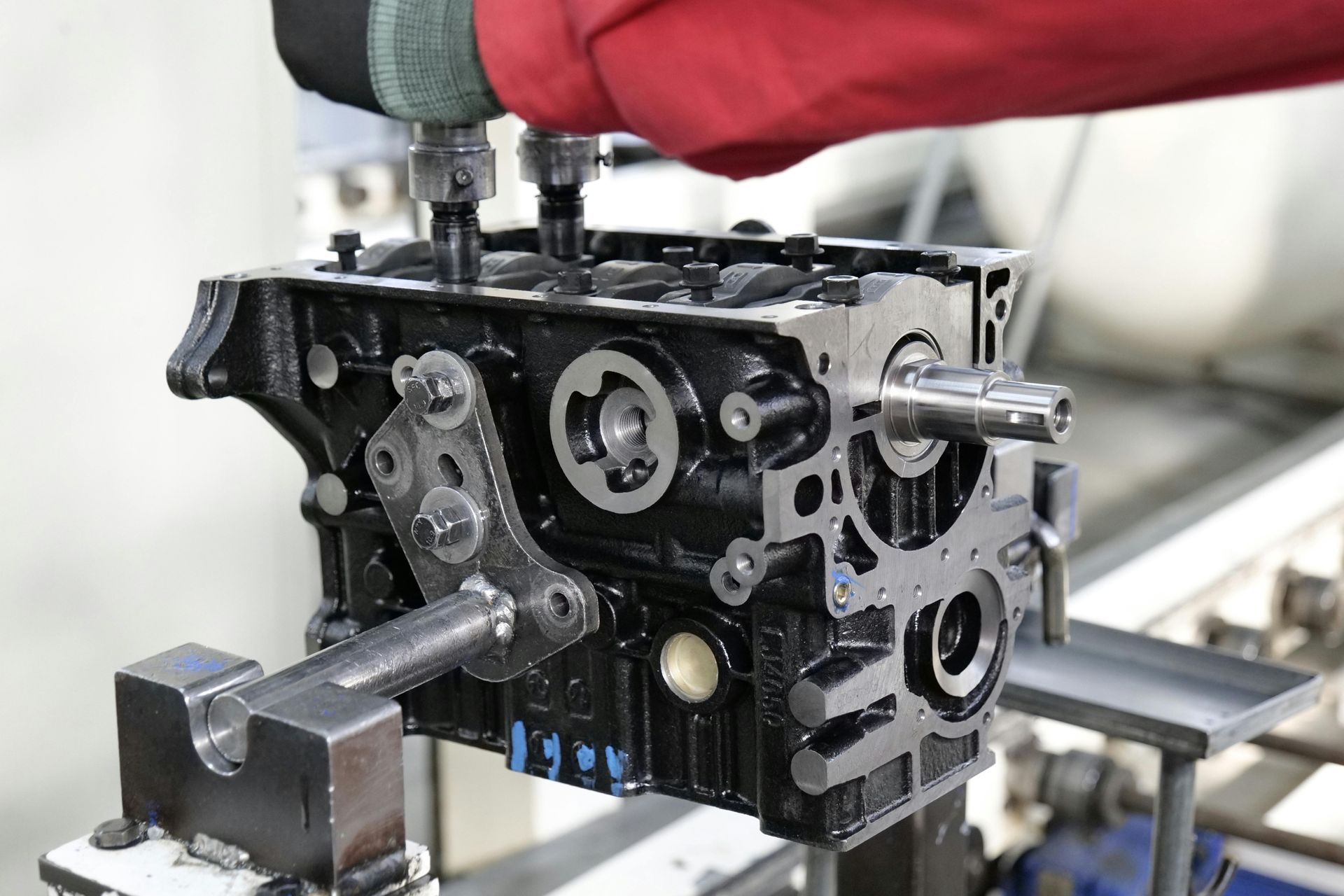 A worker in a red sleeve uses an automated tool to assemble a black automotive engine block in a factory.