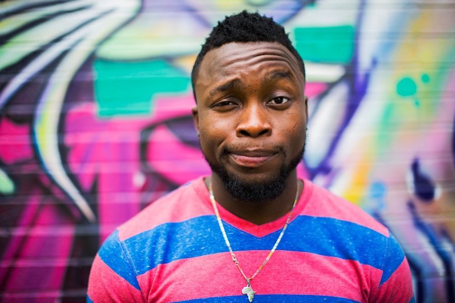 A man wearing a striped shirt and a necklace is standing in front of a graffiti wall.