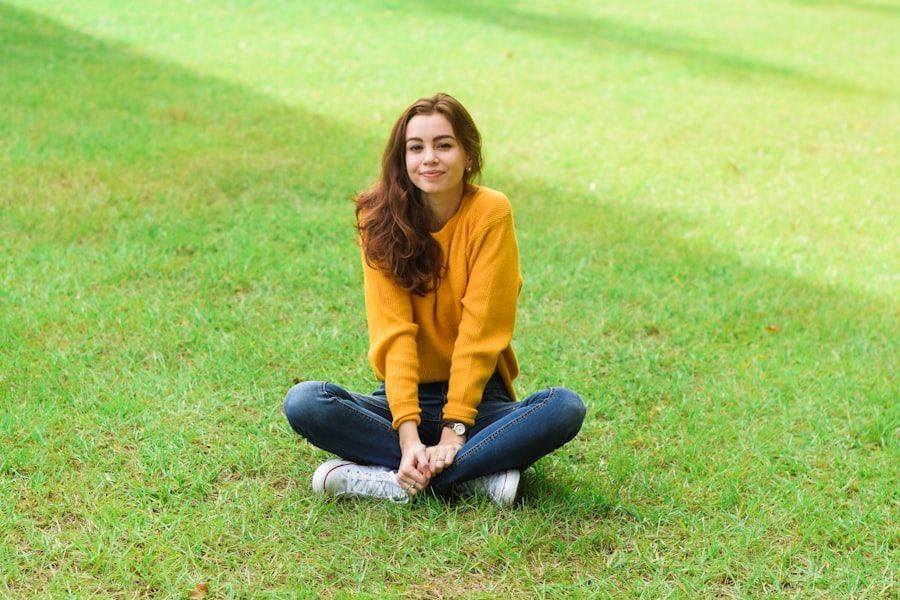 A woman in a yellow sweater is sitting on the grass with her legs crossed.