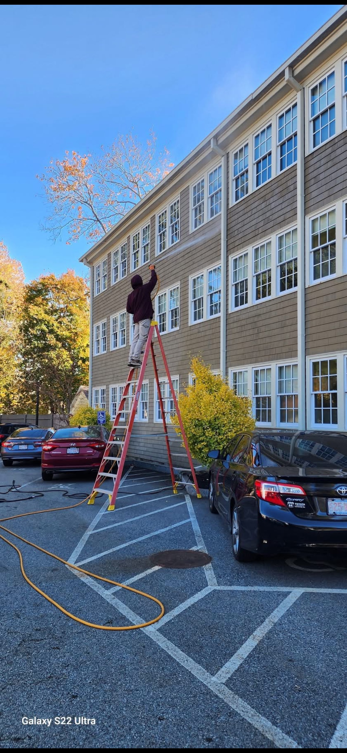 Person on a ladder cleaning a building's gutter. Cars parked in the lot, blue sky, and fall foliage.