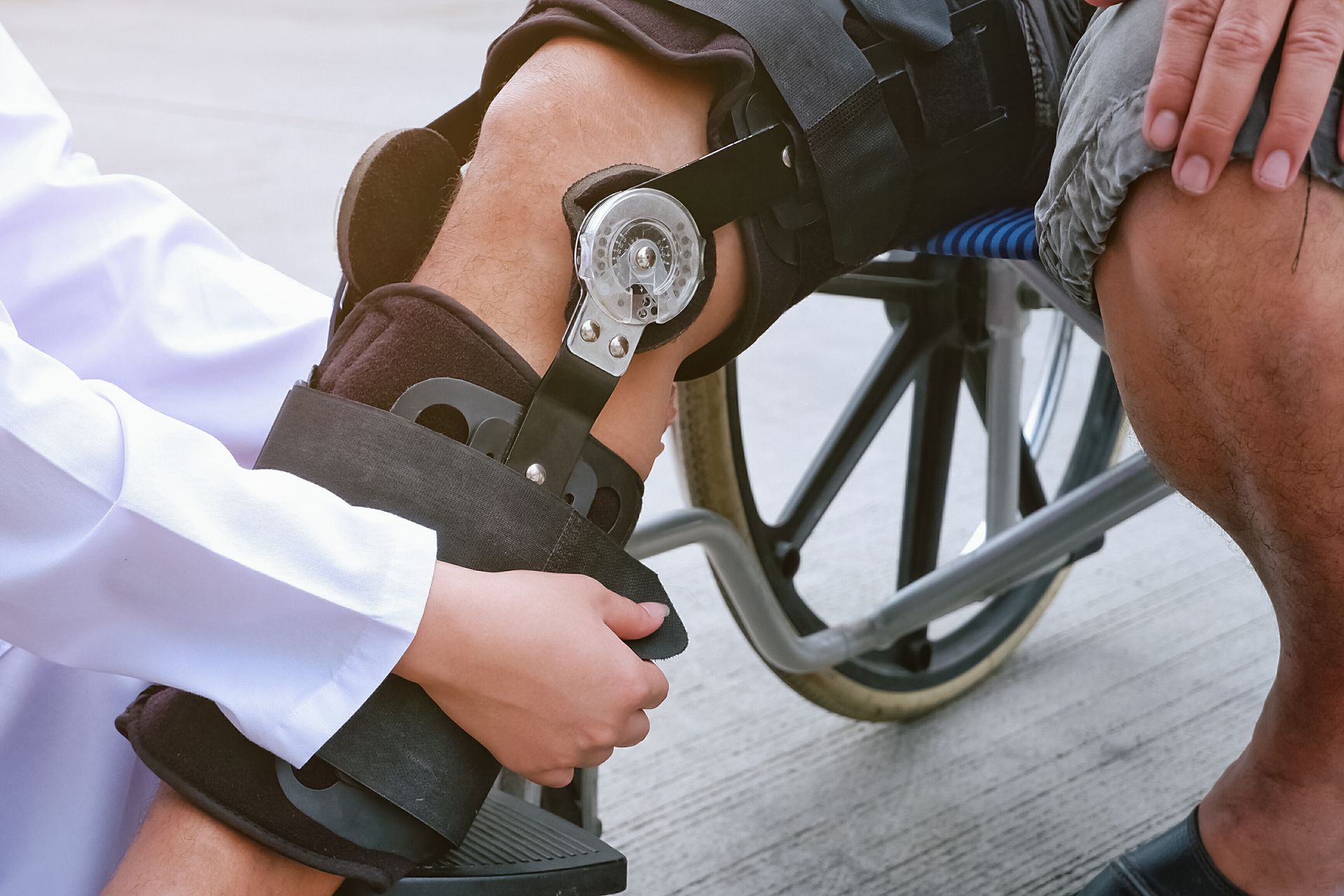 Person in wheelchair with knee brace, being adjusted by a medical professional.
