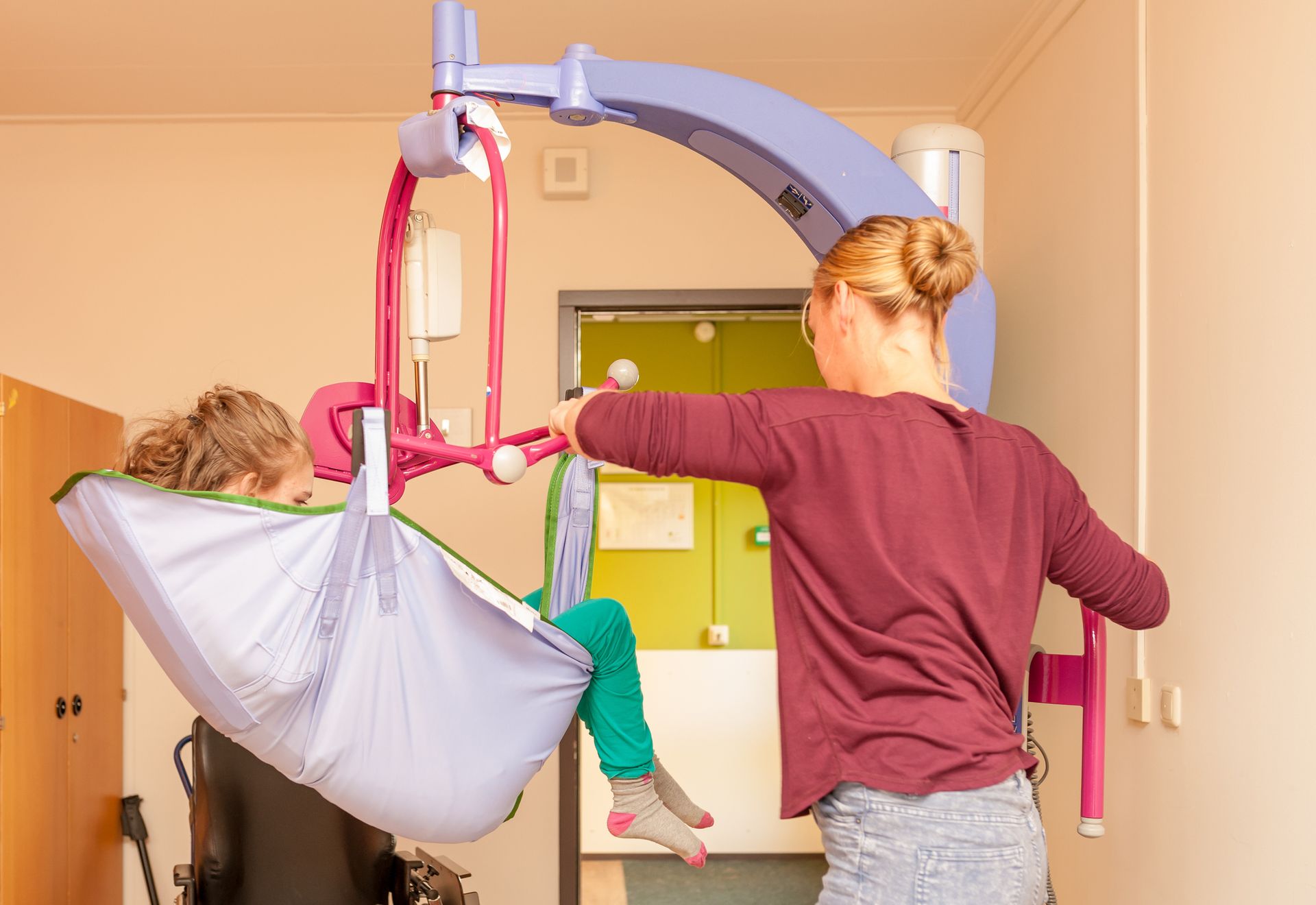 Person assisting a person in a hoist, lilac lift, green and pink sling, interior setting.