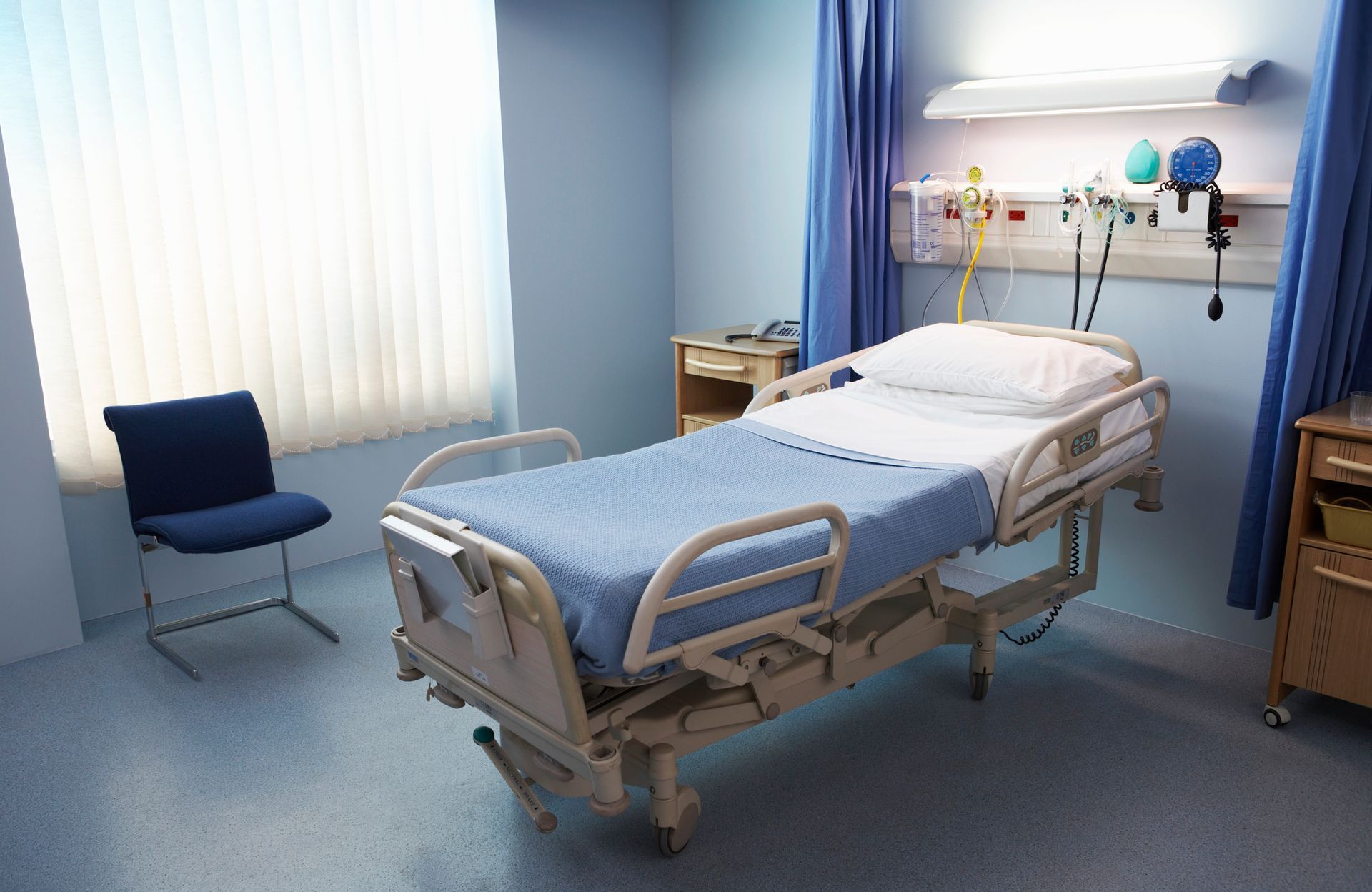 Empty hospital room with adjustable bed, blue curtains, and medical equipment.