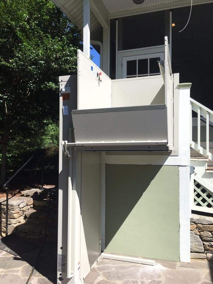Outdoor vertical platform lift next to porch. Beige, with metal platform, green wall.