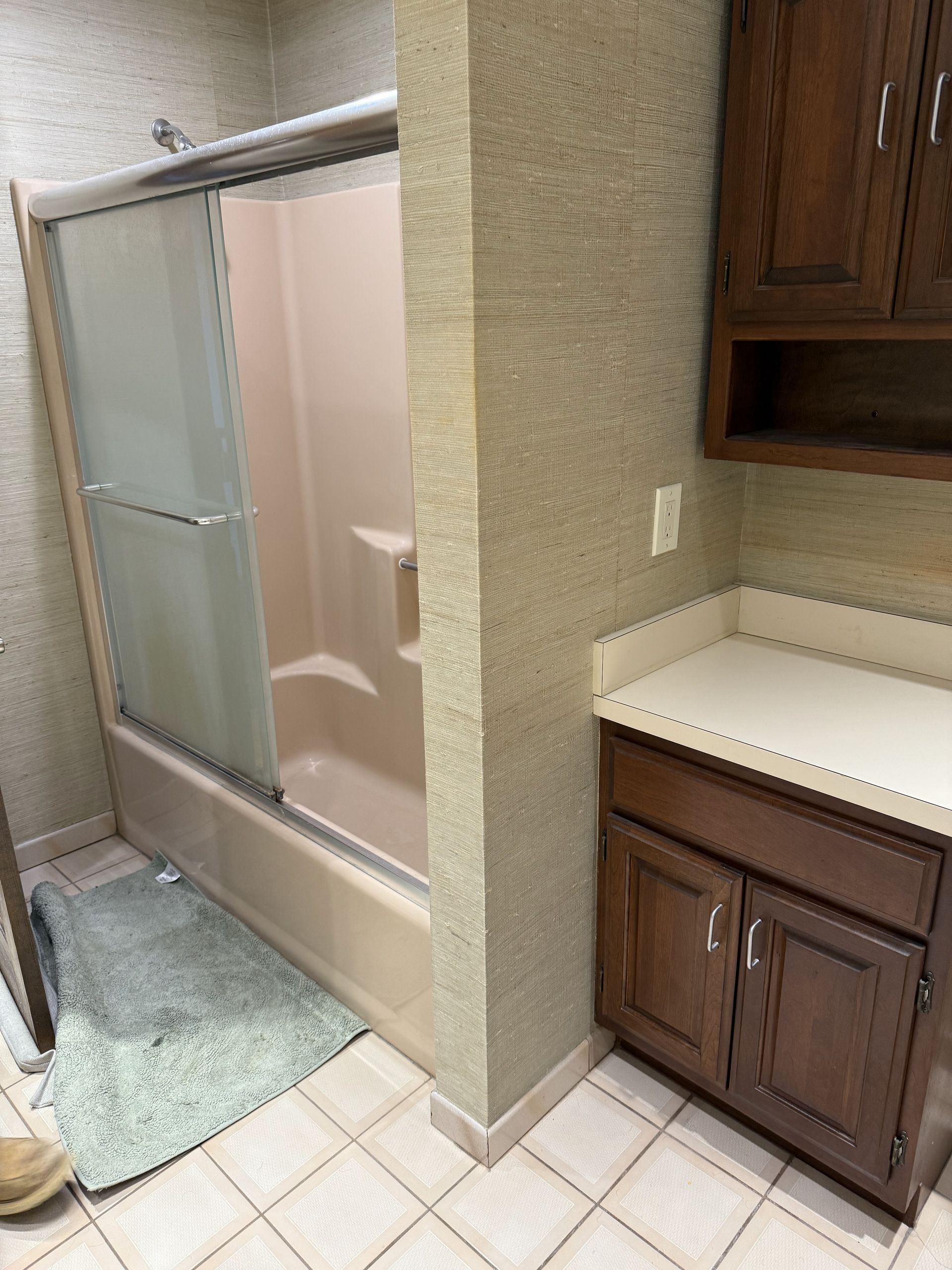 Bathroom with a pink shower/tub, vanity, and tan patterned walls. Light blue bath mat on the tiled floor.