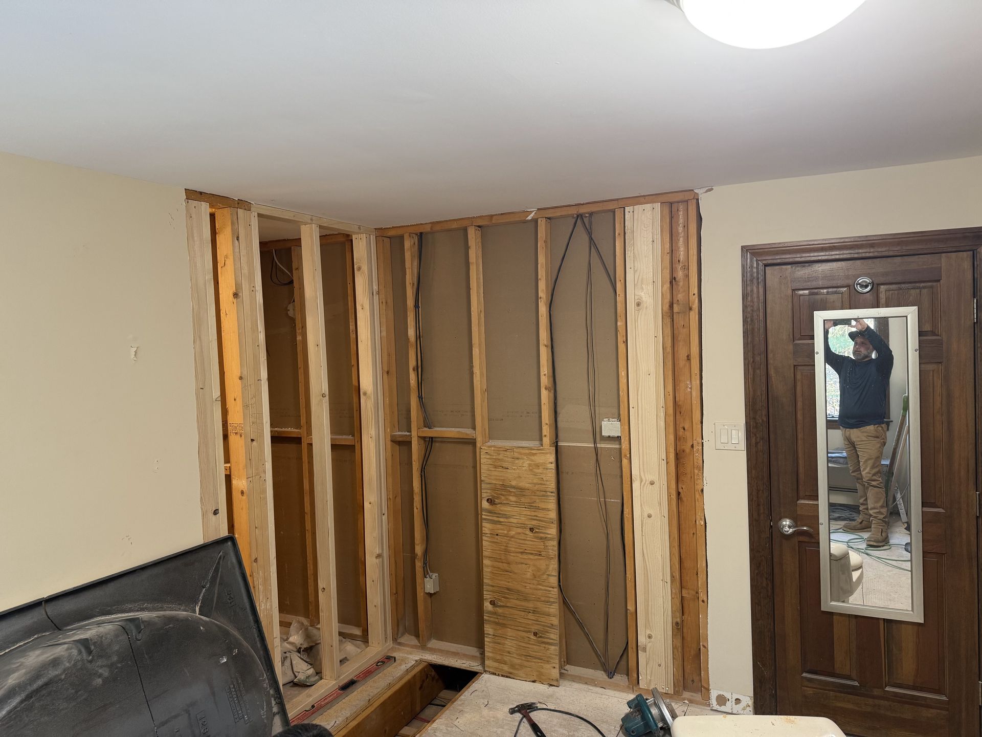 Interior wall in construction; exposed wooden frame, wires, beige wall, dark wooden door with mirror.