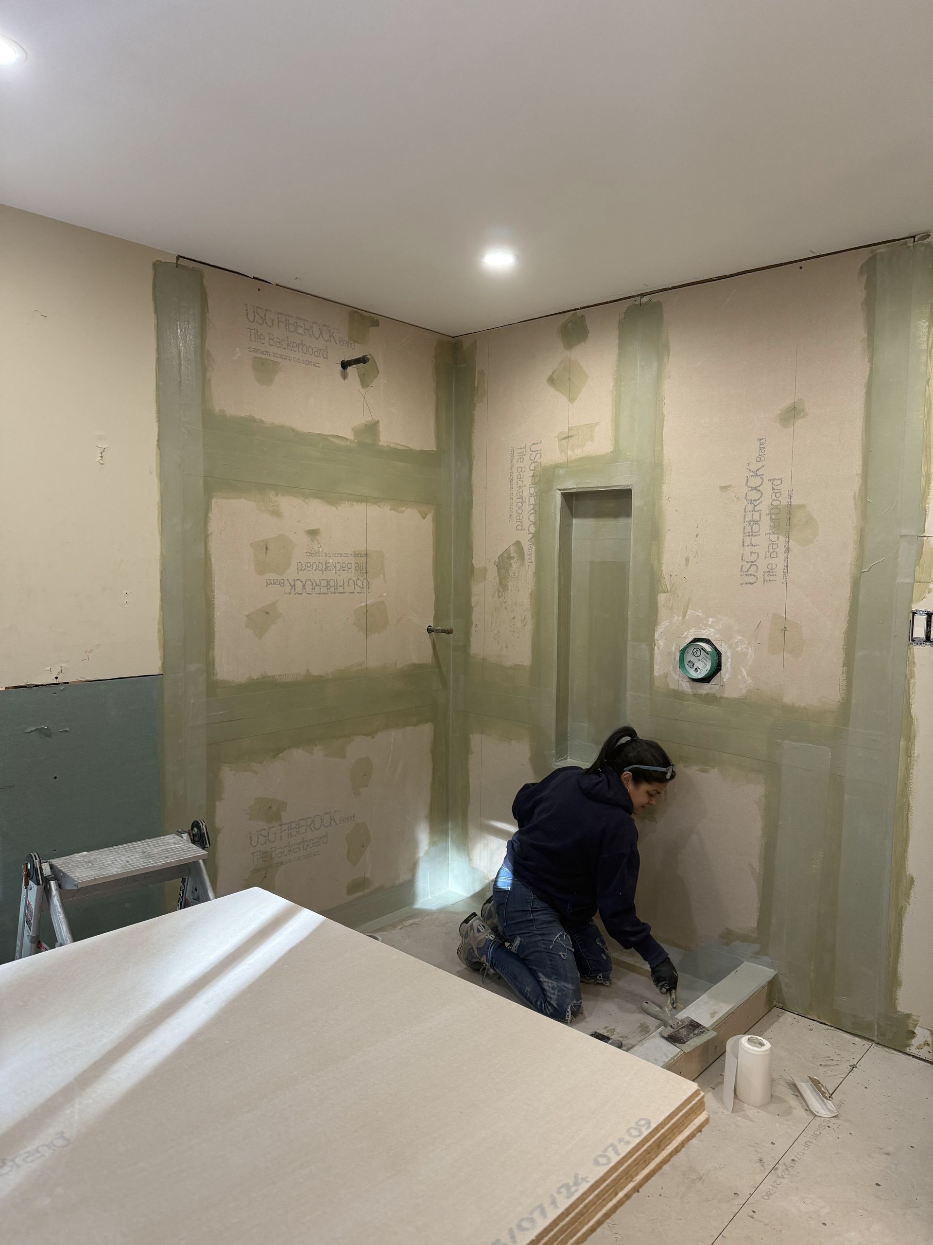 Person kneeling, spreading compound on bathroom wall during renovation. Walls are green and beige.