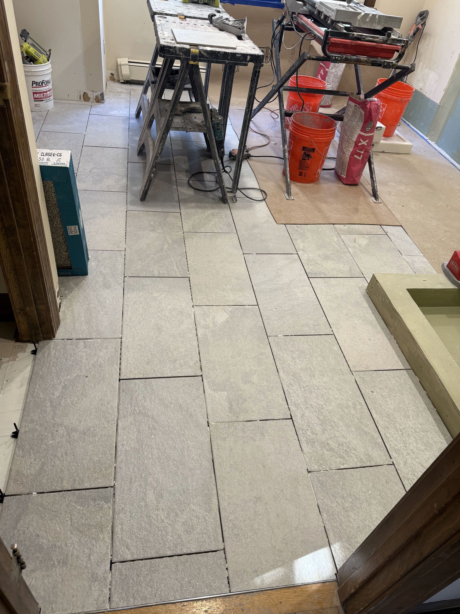 Gray floor tiles partially installed in a room; tools and buckets are visible in the background.