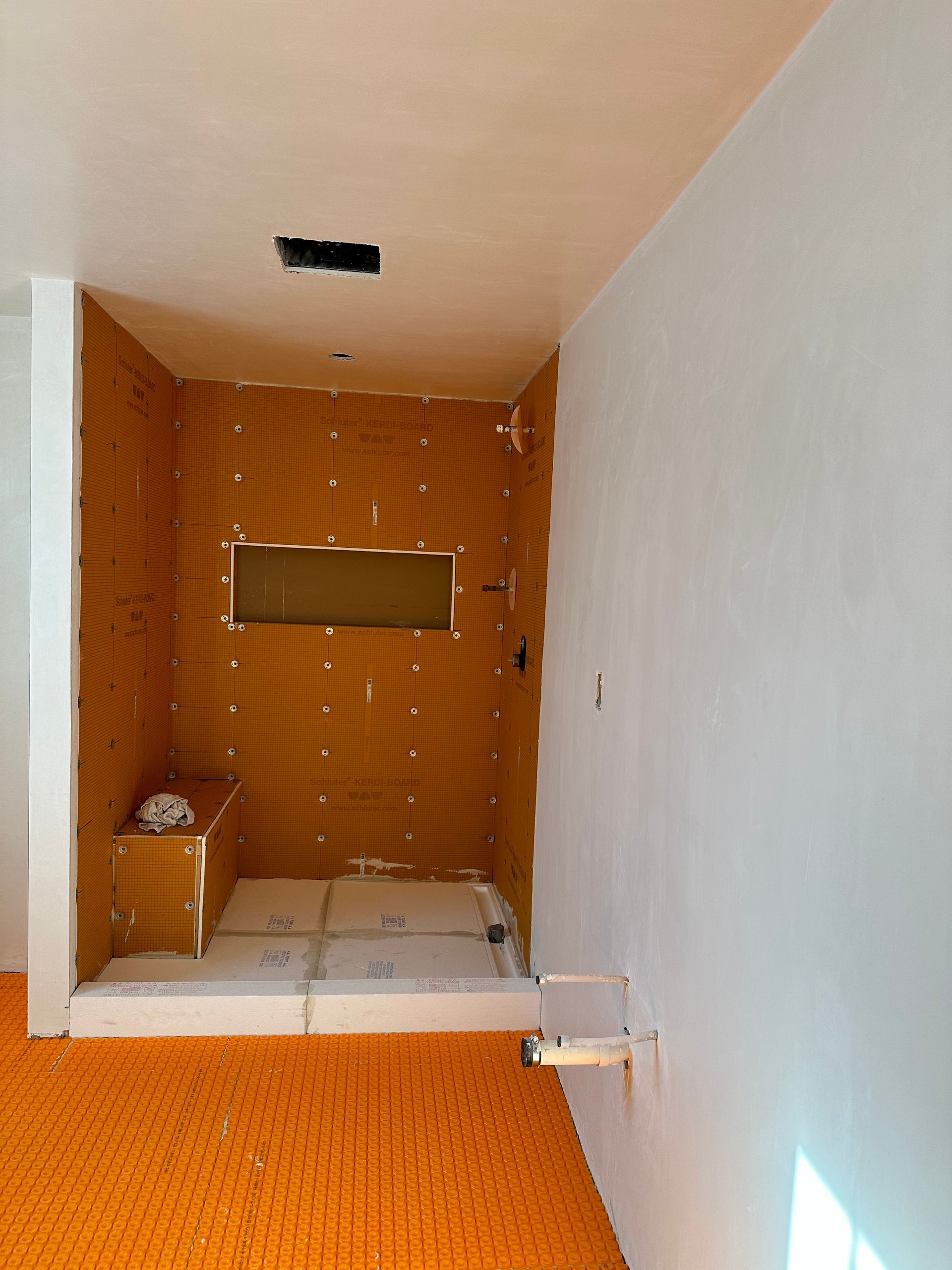 Bathroom remodel in progress; orange waterproof board walls, white walls/ceiling, bench, plumbing visible.