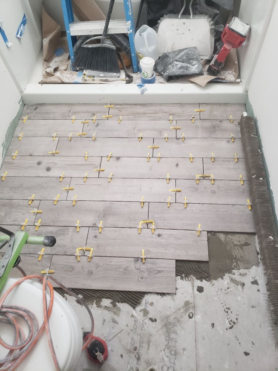 Bathroom floor with wood-look tiles being installed. Yellow spacers are between tiles, tools and ladder are visible.