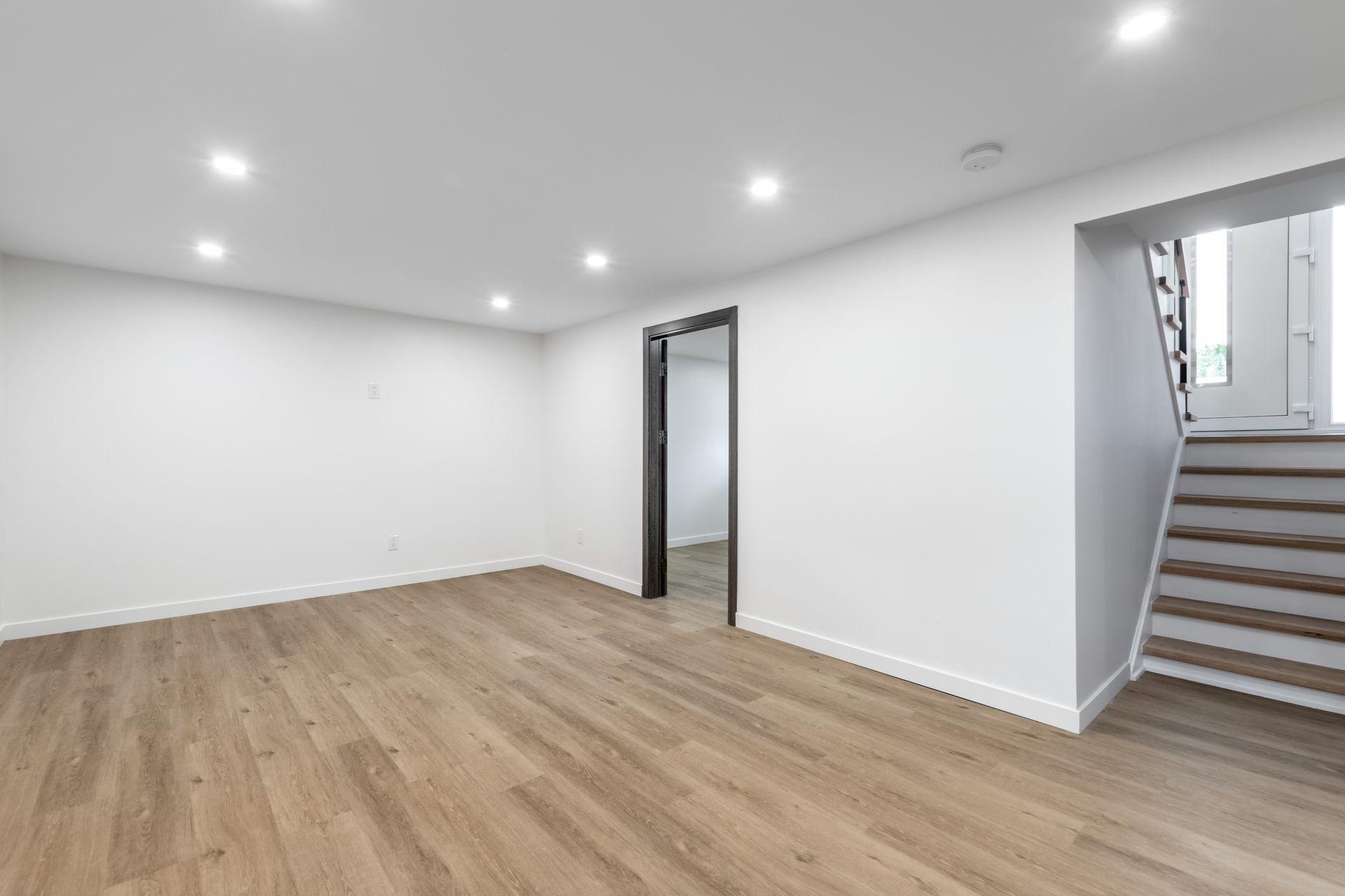Empty room with wood floors, white walls, recessed lights, and a staircase.
