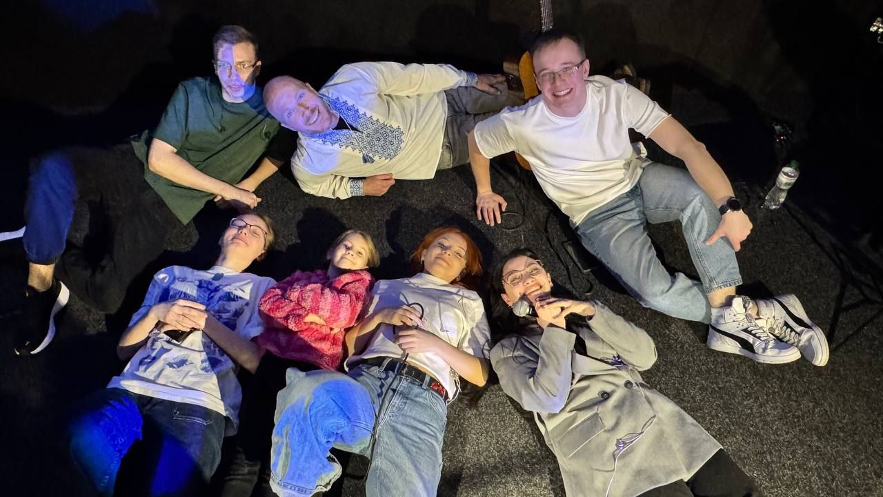 Gruppe på sju personer smiler og ligger på gulvet under scenebelysning.