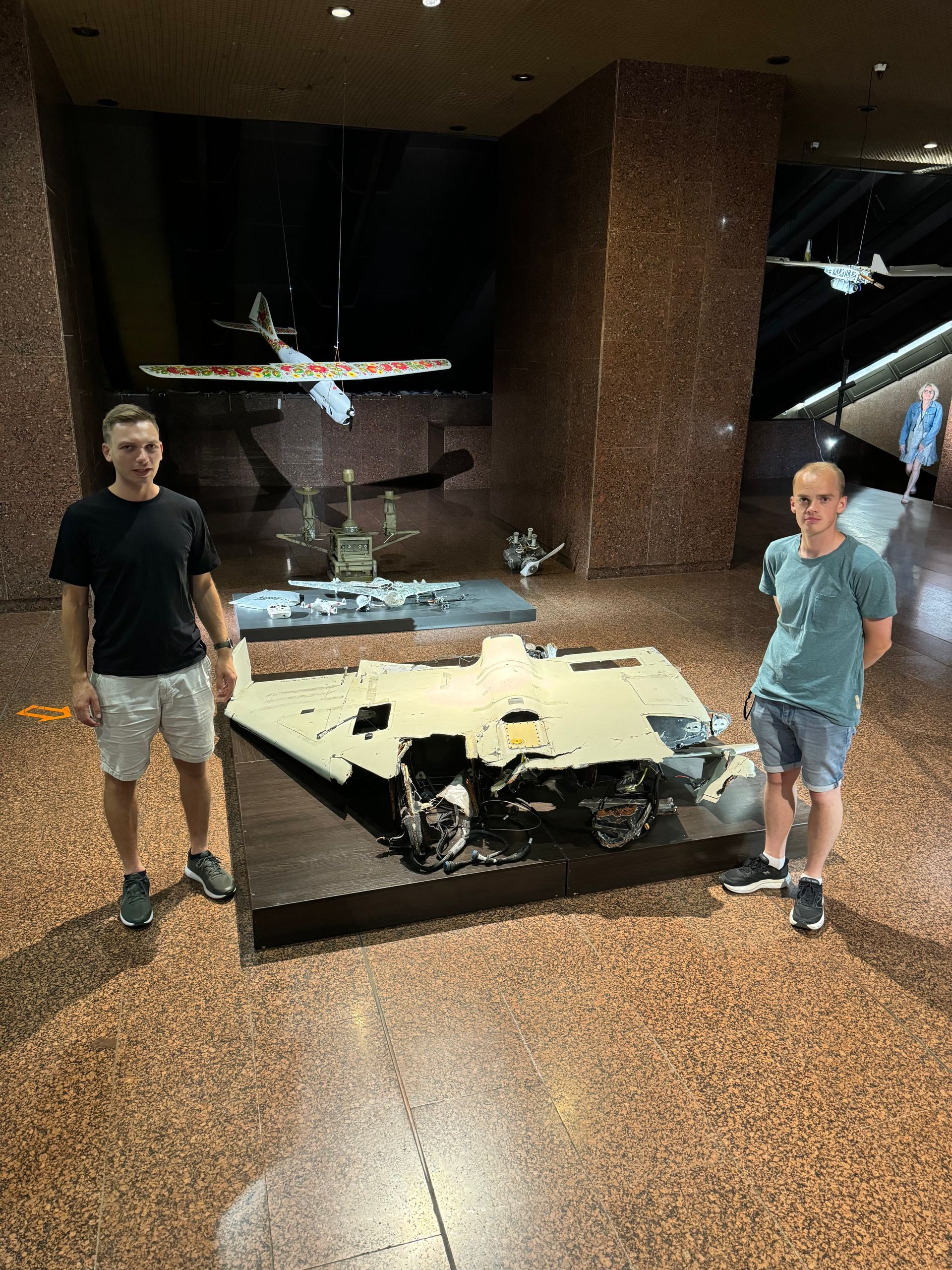To personer står ved en vrakdelsutstilling på et museum. Utstillingen er brun og grå, med et fly over hodet.