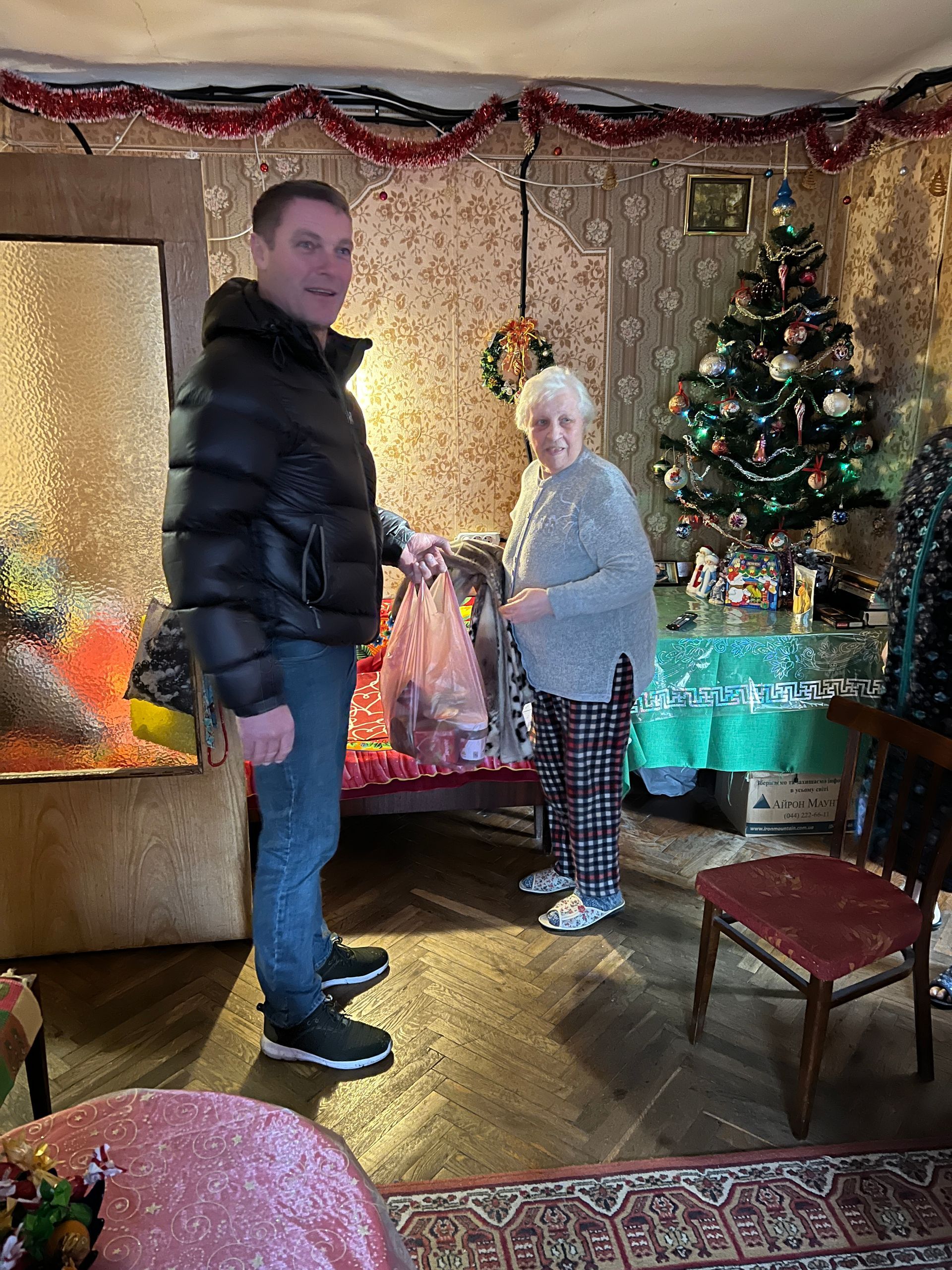 Mann gir en gavepose til en eldre kvinne i et pyntet rom med et juletre.