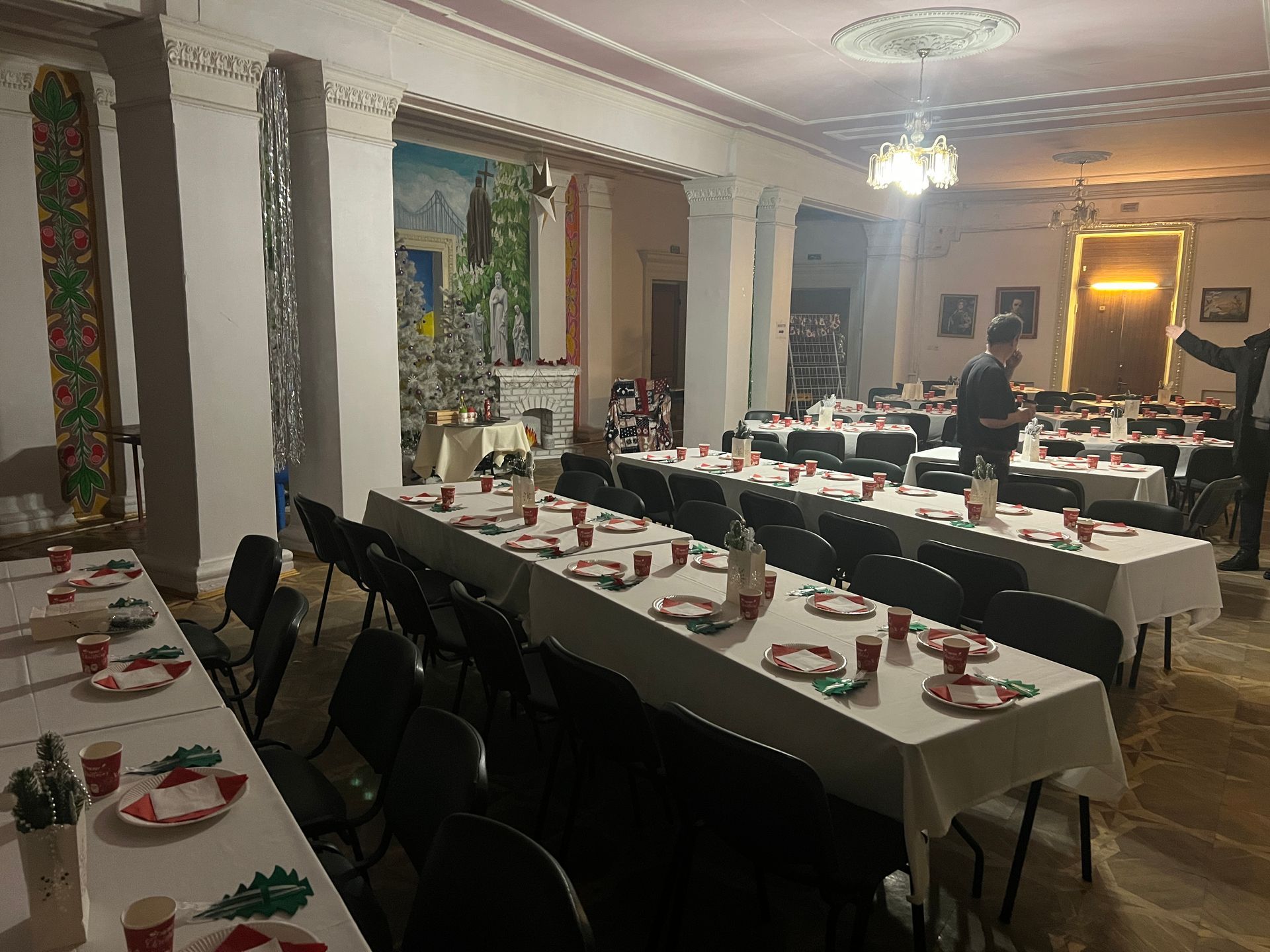En festsal med lange bord dekket for et måltid. Folk står ved bordene, noen forbereder seg.