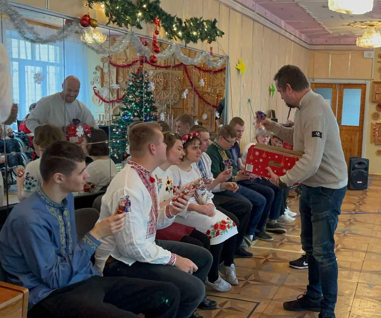Mann gir gaver til en sittende gruppe. De er i et pyntet rom med et juletre. Noen har på seg broderte skjorter.