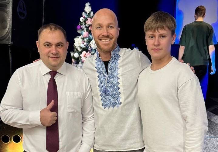 Tre menn som smiler og poserer for et bilde, en av dem har på seg en hvit skjorte med blå broderi, med et pyntet juletre i bakgrunnen.