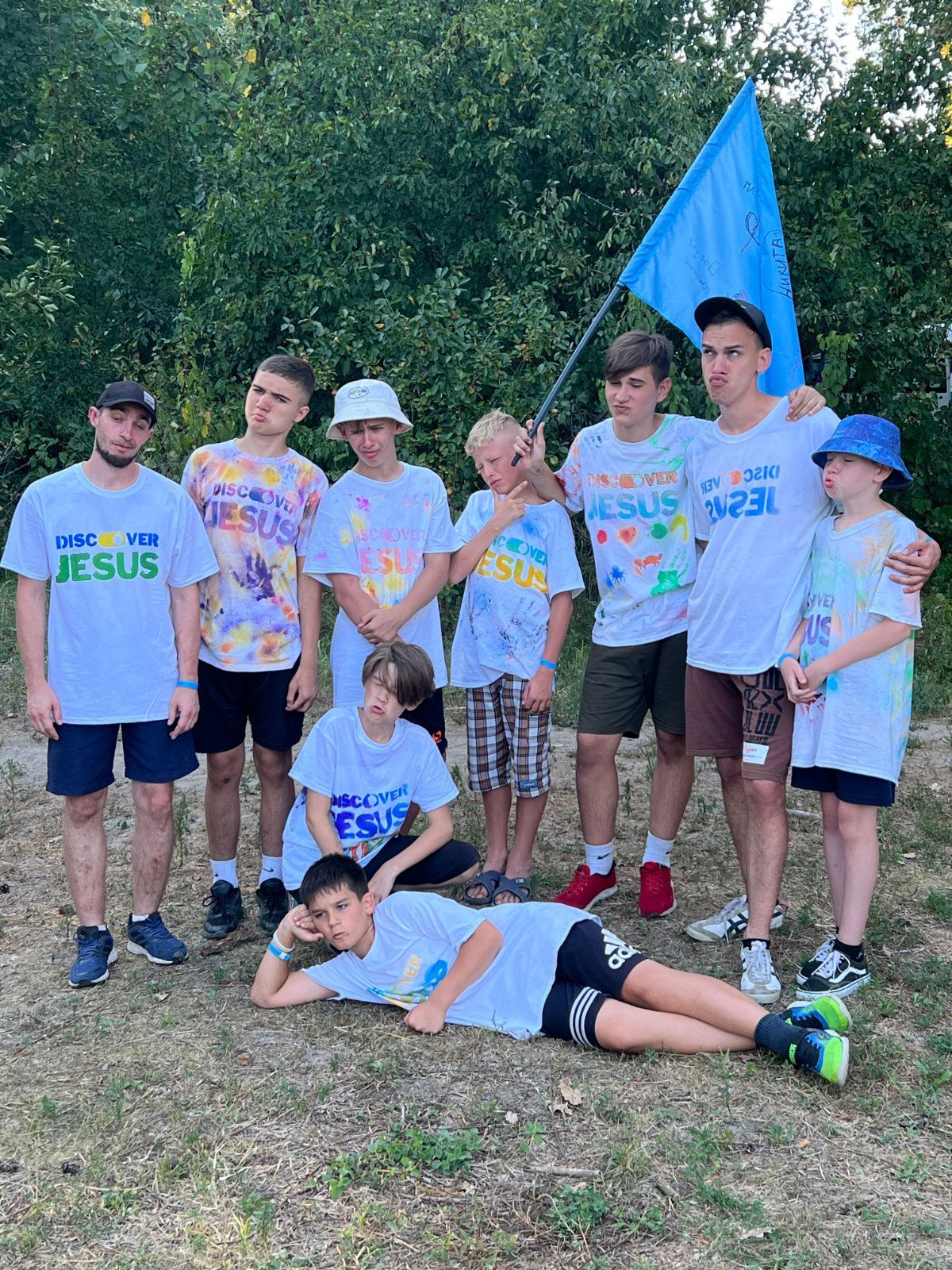 Gruppe mennesker i hvite skjorter med teksten «Oppdag Jesus», som holder et blått flagg utendørs.Dare2be.org arrangerer sommerleir i Ukraina med Helge Flatøy