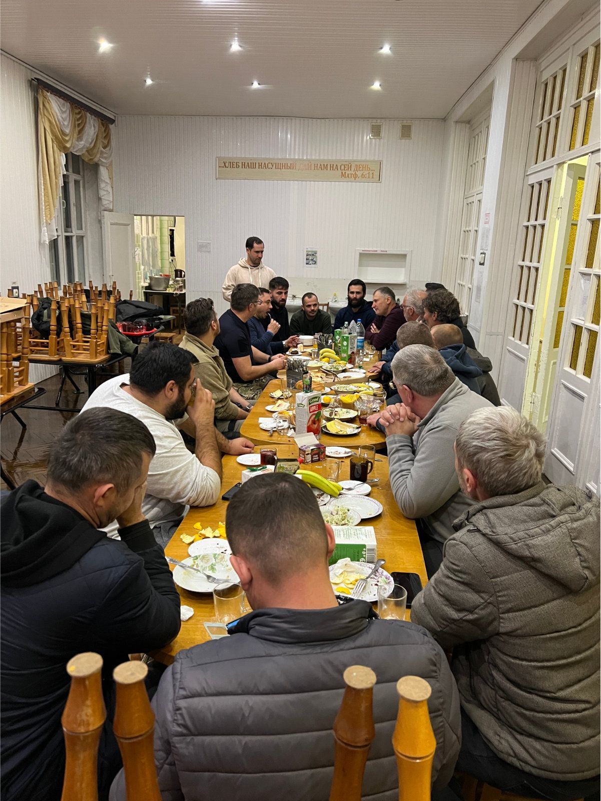 Gruppe mennesker sitter rundt et bord, muligens på en sammenkomst eller et måltid i et sterkt opplyst rom.