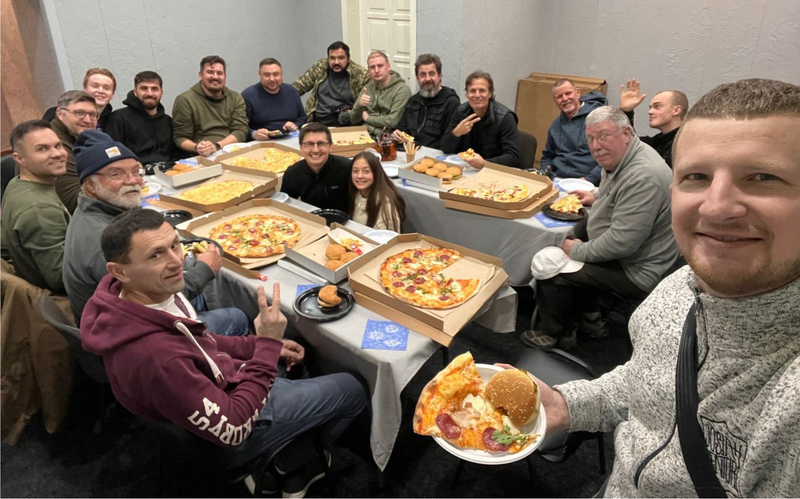 Gruppe mennesker sitter rundt et bord og koser seg med pizza. Noen holder en tallerken med mat. Rominnredning.