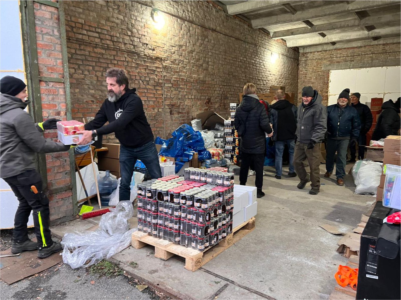 Folk mottar forsyninger fra frivillige ved en lastebrygge; esker og paller med varer.