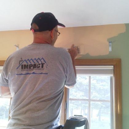 Person painting a beige wall above a window in a room, wearing a black cap and gray shirt