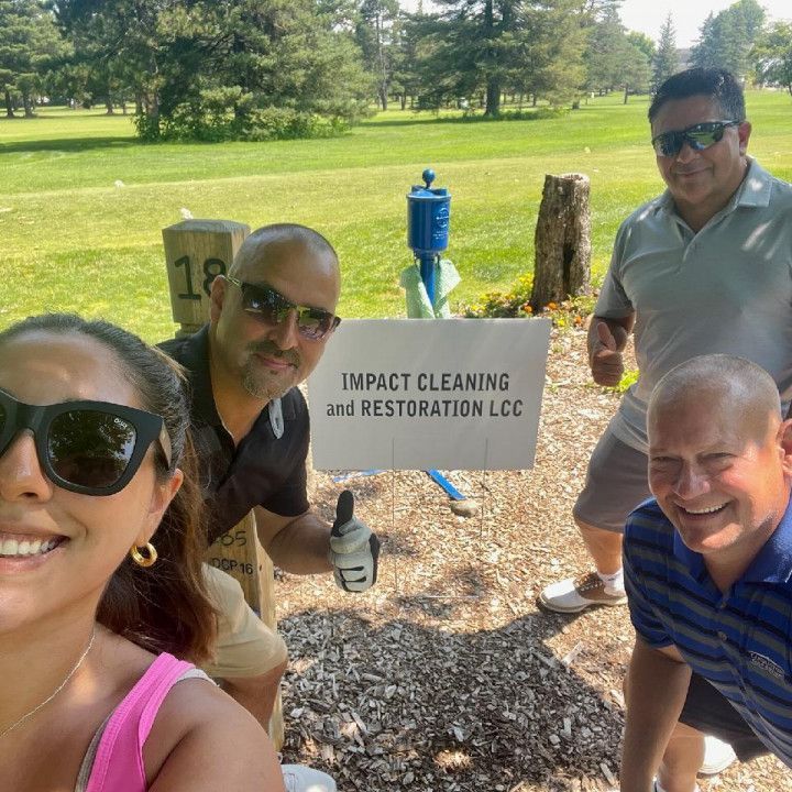 Four people smiling by an “Impact Cleaning and Restoration LLC” sign in a sunny park