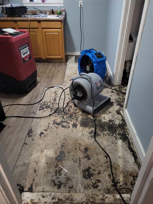 Damaged kitchen floor with water cleanup fans, cords, and debris beside cabinets and a wall.