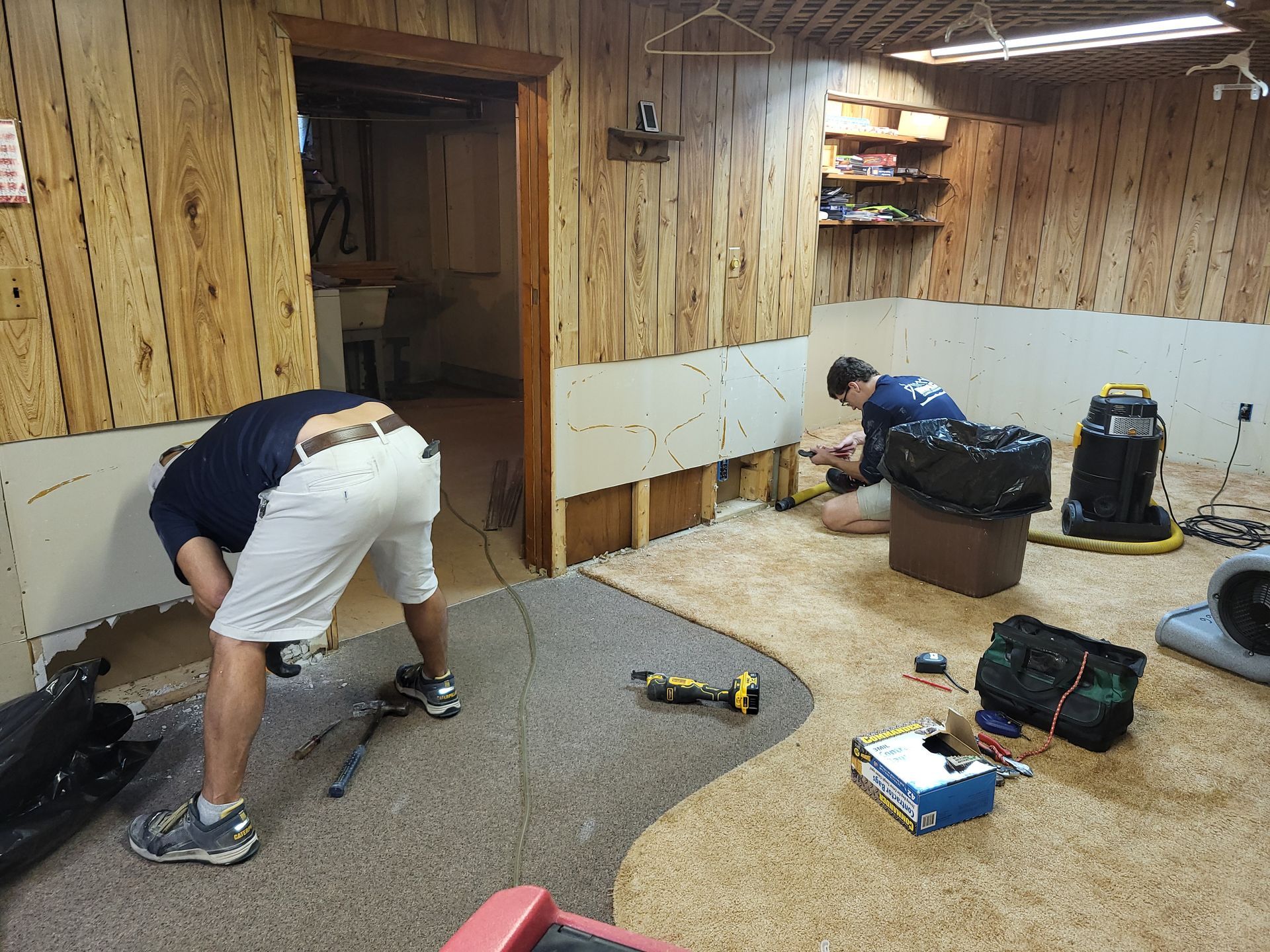 Two people cleaning and organizing a wood-paneled room with tools and supplies on the floor