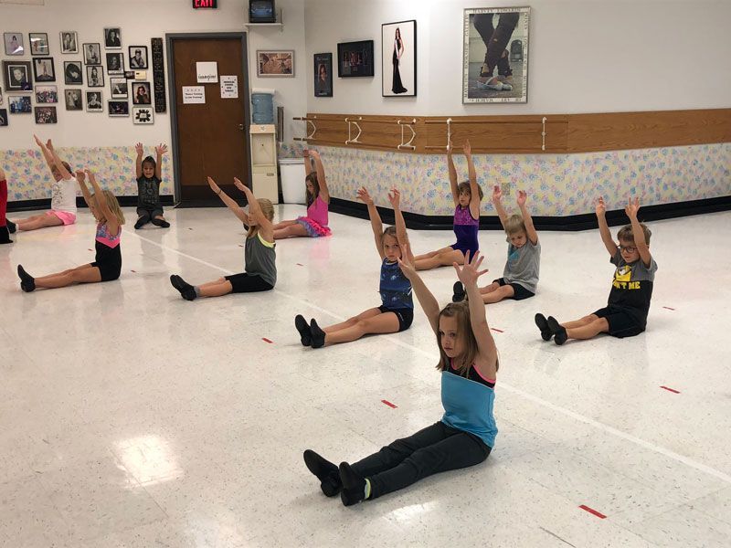 Children stretching on the floor in a dance studio, arms raised in a seated split position.