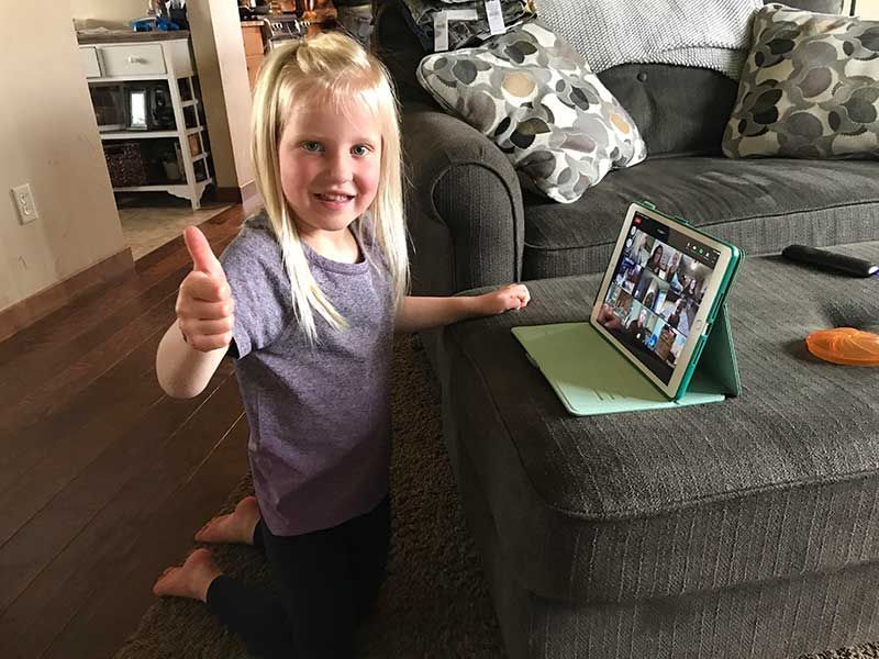 Child kneeling beside a couch, smiling and giving a thumbs-up next to a tablet on a green stand.