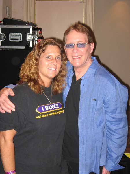 Two people smiling and posing together indoors, one in a black T-shirt and the other in a blue shirt and sunglasses.