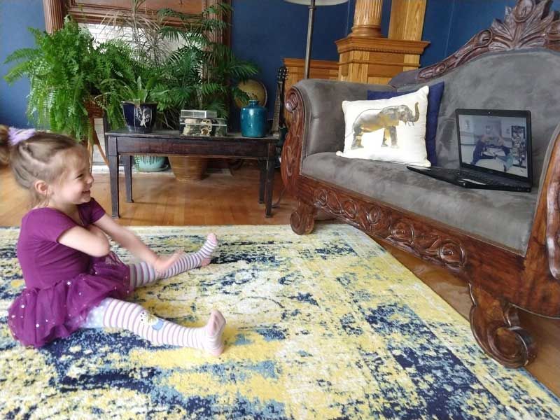 Child sitting on a patterned rug in a living room, facing a couch with a laptop and pillow