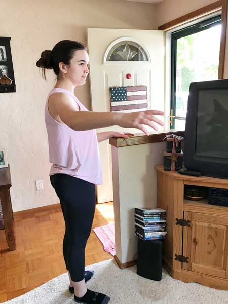 Woman standing with arms extended toward a TV in a living room near a window.