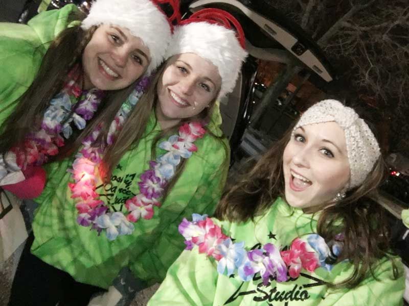 Three smiling people in Santa hats and green outfits, wearing leis, posing outdoors at night.
