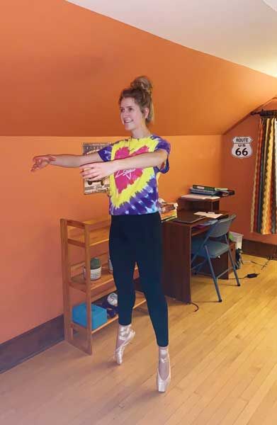 Person standing on pointe in a room with an orange wall, arms extended in a ballet pose