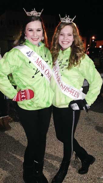 Two women in green jackets and crowns with sashes, standing together outdoors at night.